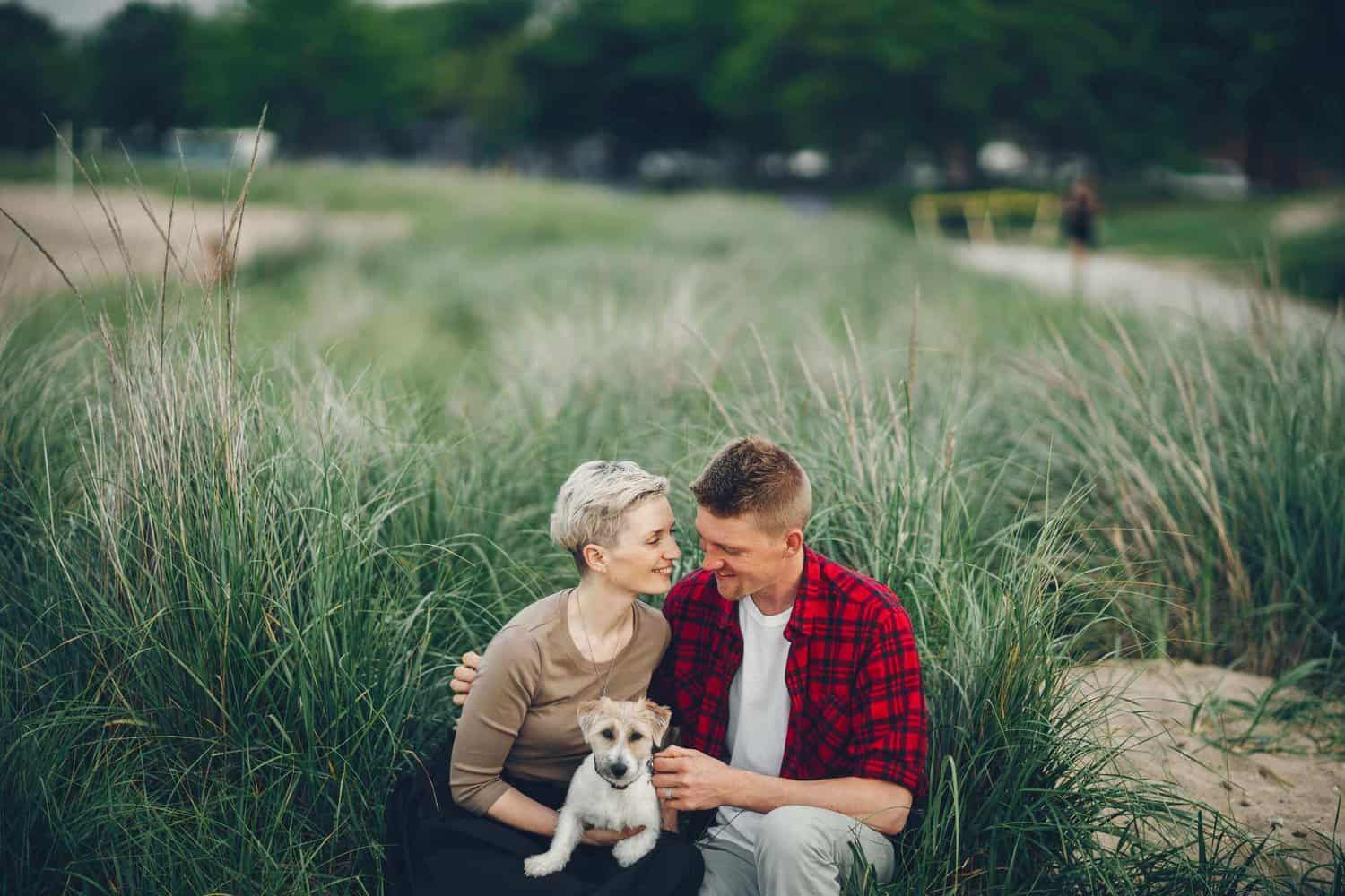 A couple stting in front of a green field holding their small dog as they gaze lovingly into each others eyes