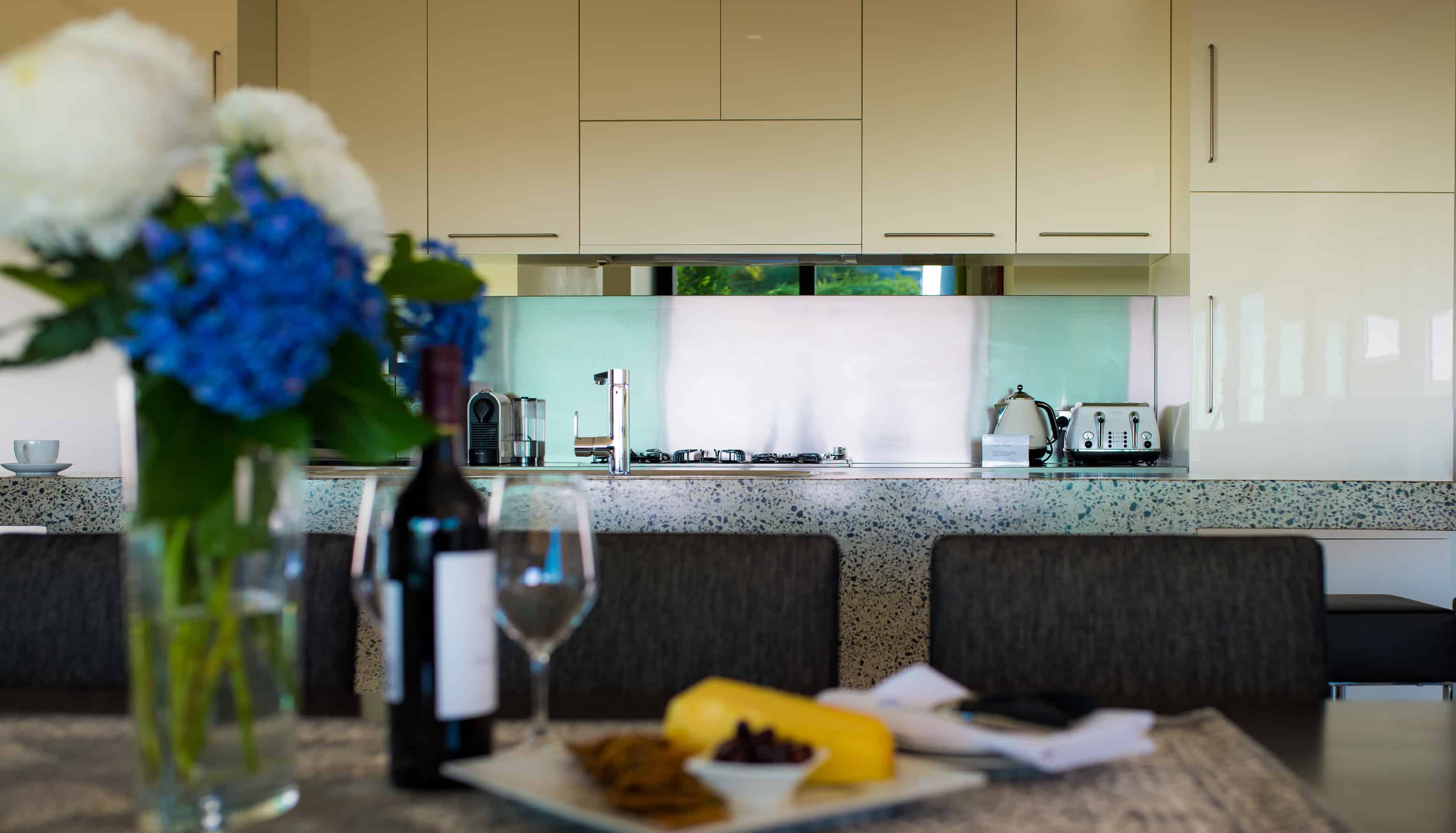 Modern kitchen with clean lines and neutral tones, featuring a bouquet of blue hydrangeas in the foreground, and a bottle of wine with glasses ready to serve.