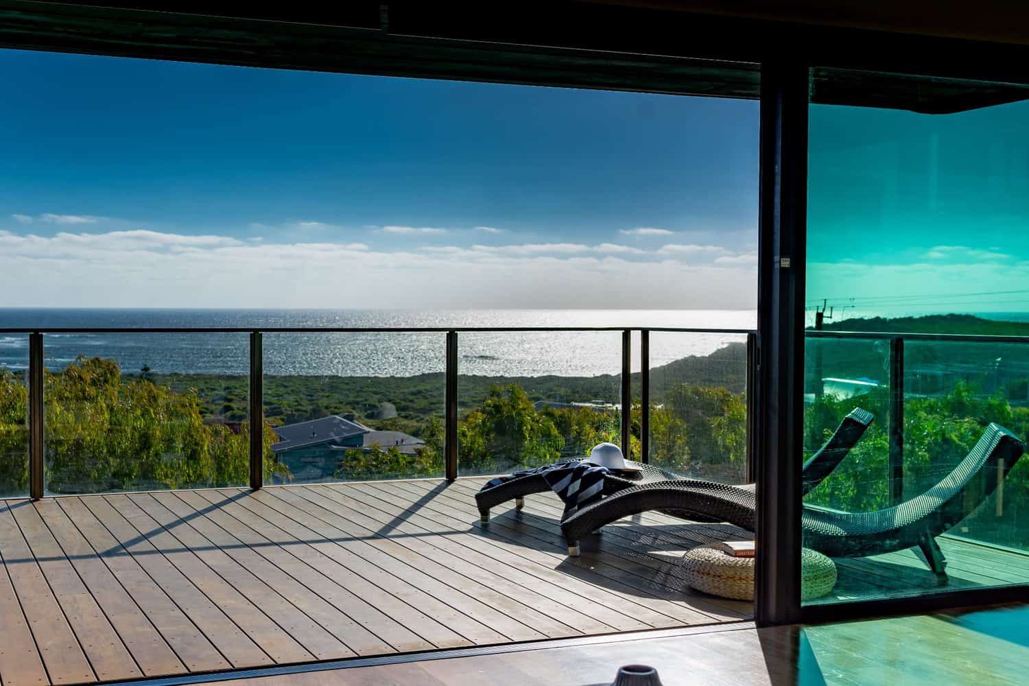 View from a seaside balcony with glass balustrades, showcasing a relaxing sun lounger overlooking the shimmering ocean under a clear sky.
