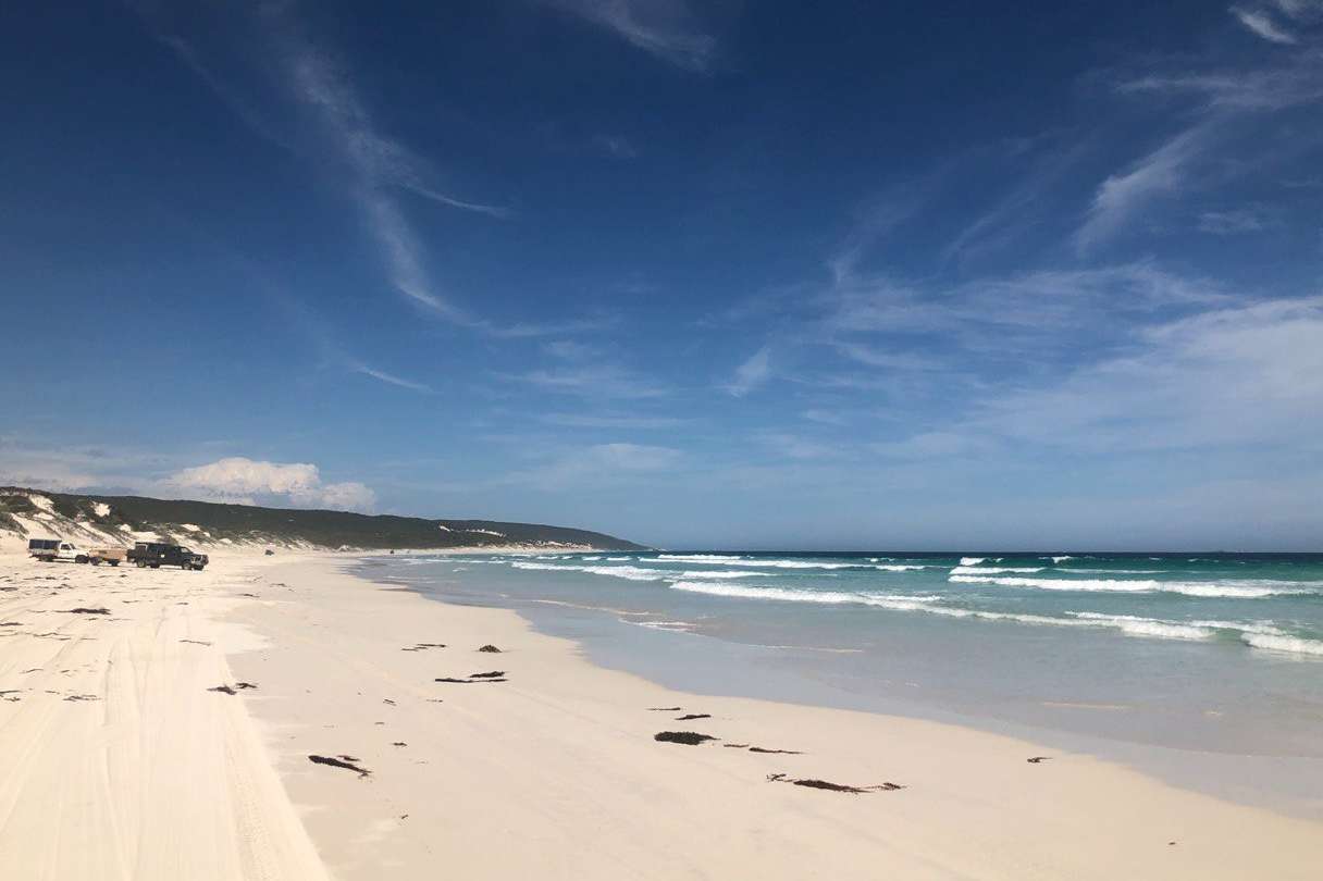 Vehicles parked on the soft white sands of an Deepdene Beach offer a sense of adventure, with the expansive shoreline leading into the distance, the ocean's waves gently breaking, and a sky painted with wispy clouds.