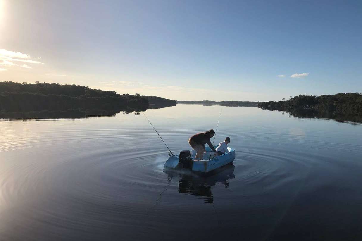 Two people on a small boat fishing on the calm waters of the Blackwood River in Augusta at dusk, with a serene reflection of the forest-lined shore and a clear sky enhancing the peaceful fishing experience.