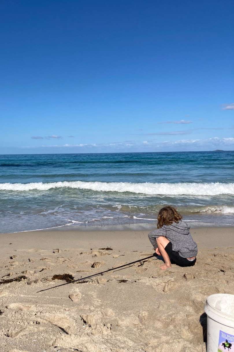 A child crouches on the sandy shore at Margaret River, attentively watching a fishing line in the surf, with a pristine blue ocean and sky backdrop.