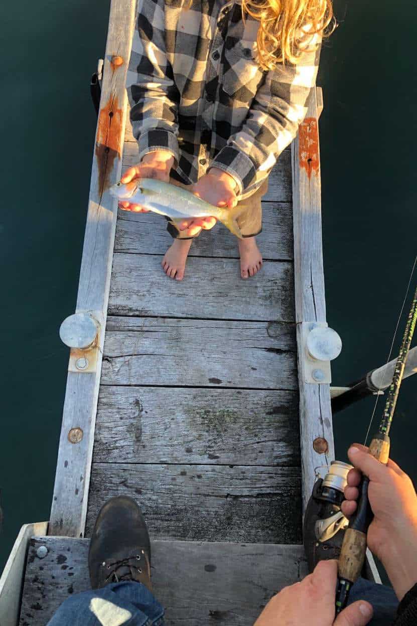 Top-down view of a child holding a small fish on a wooden jetty, with an adult's hand holding a fishing rod, capturing a family moment of a successful catch at Margaret River.