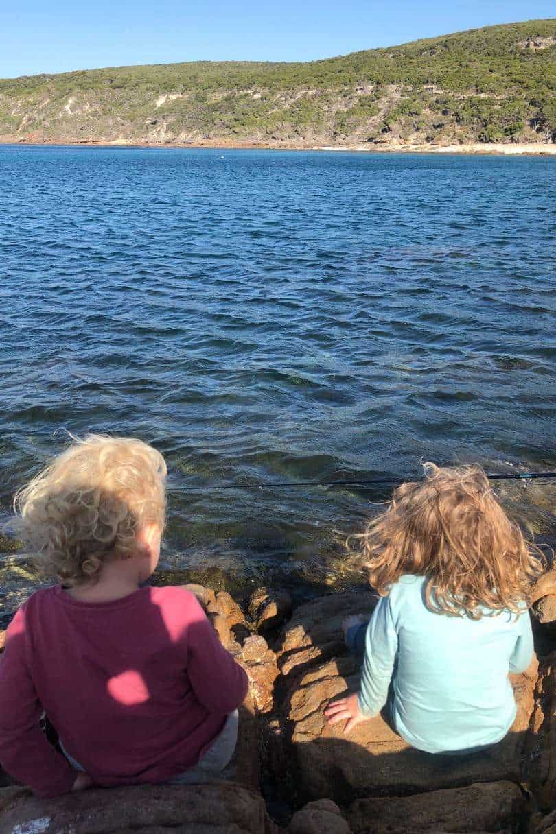 Two children sit on rocky terrain by the calm waters of Canal Rocks, with a hill in the distance, creating a peaceful scene that may inspire family fishing trips.