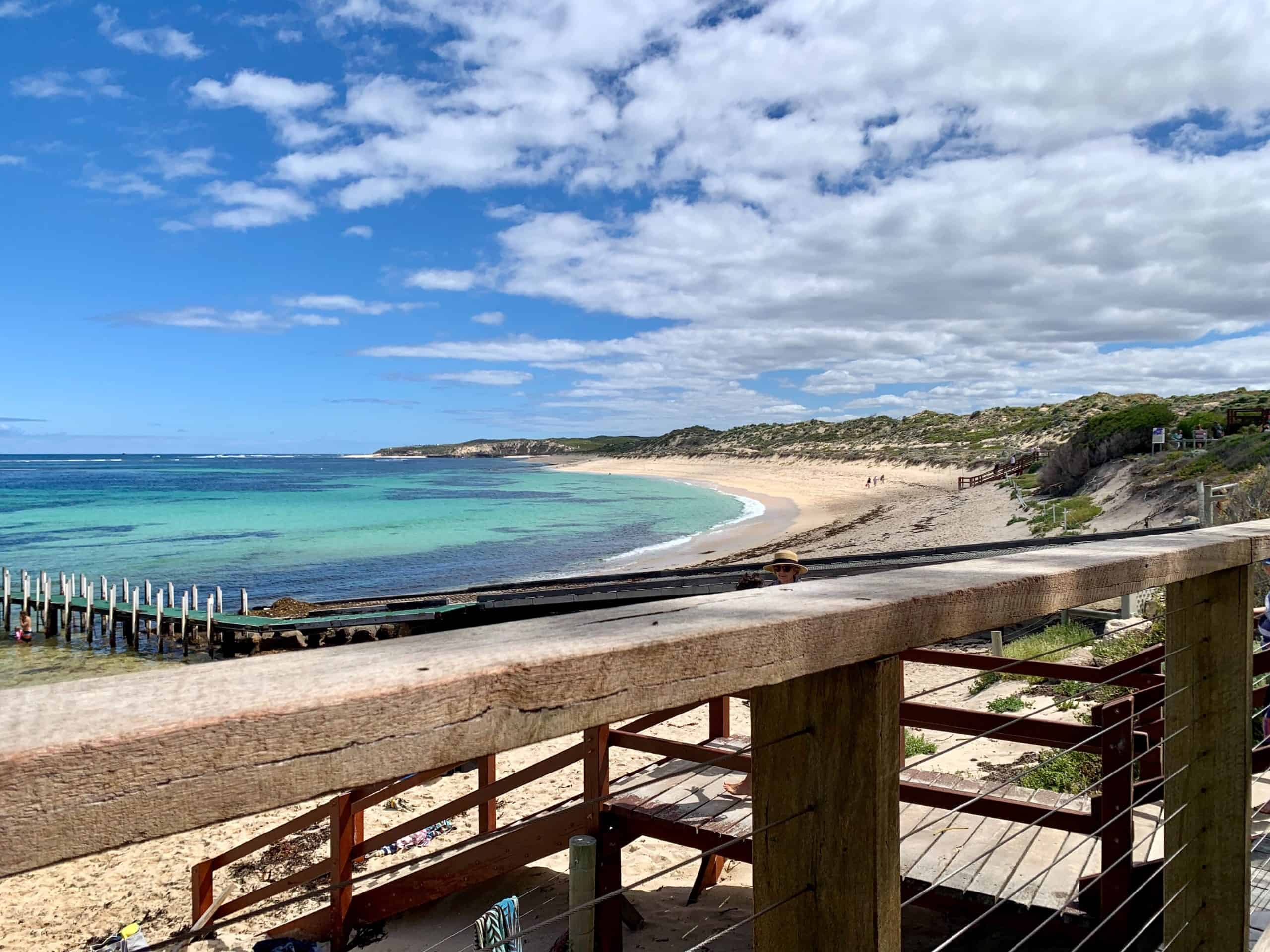 View from a wooden deck framing the stunning curve of a Margaret River beach, where crystal-clear turquoise waters meet a sandy shore, with a rustic jetty extending into the sea and sand dunes under a partly cloudy sky.