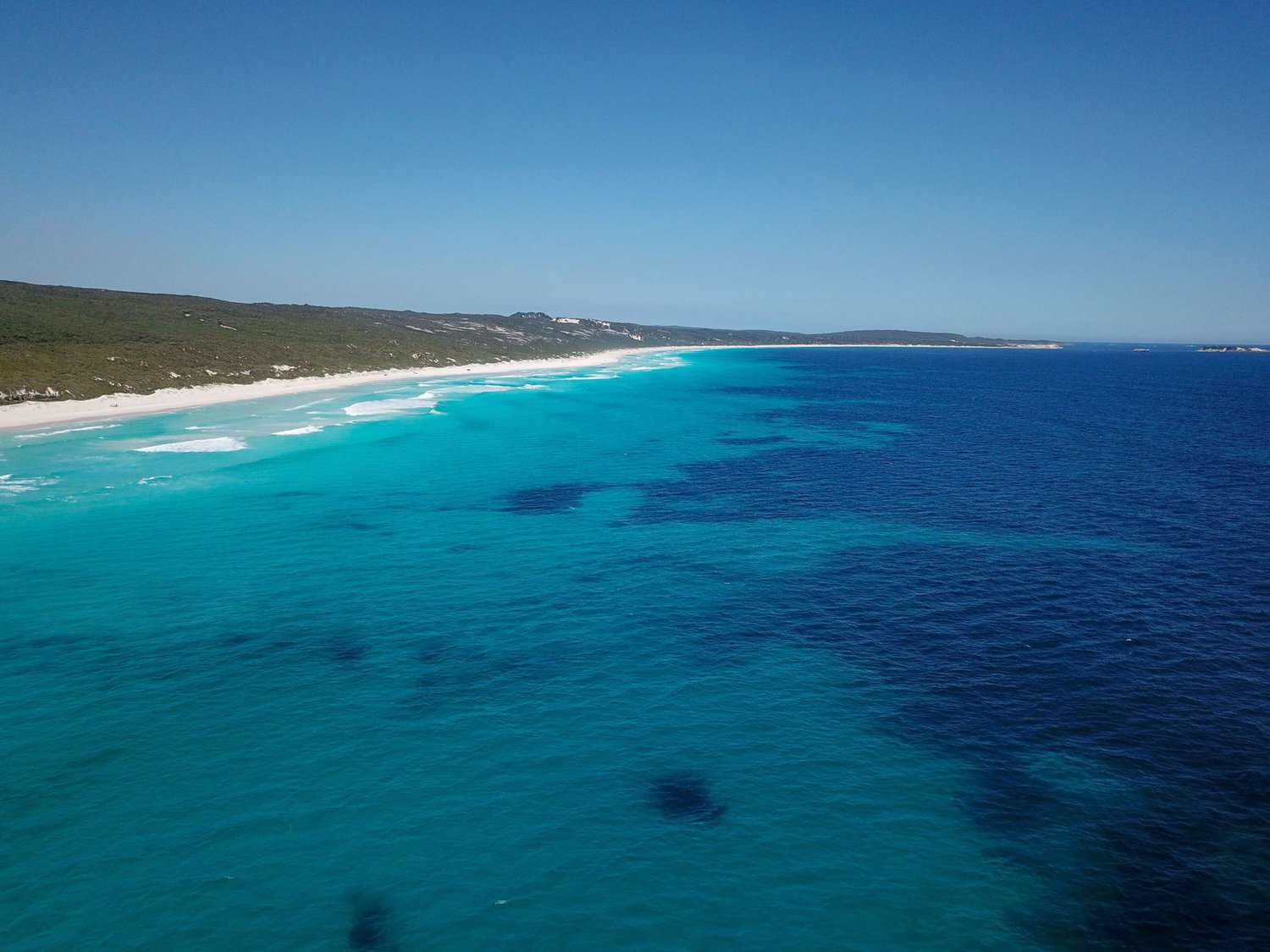 An aerial view of the serene and expansive Augusta beaches, where the brilliant turquoise of the Indian Ocean meets the pristine white sands of the coastline, bordered by lush greenery under a clear blue sky.