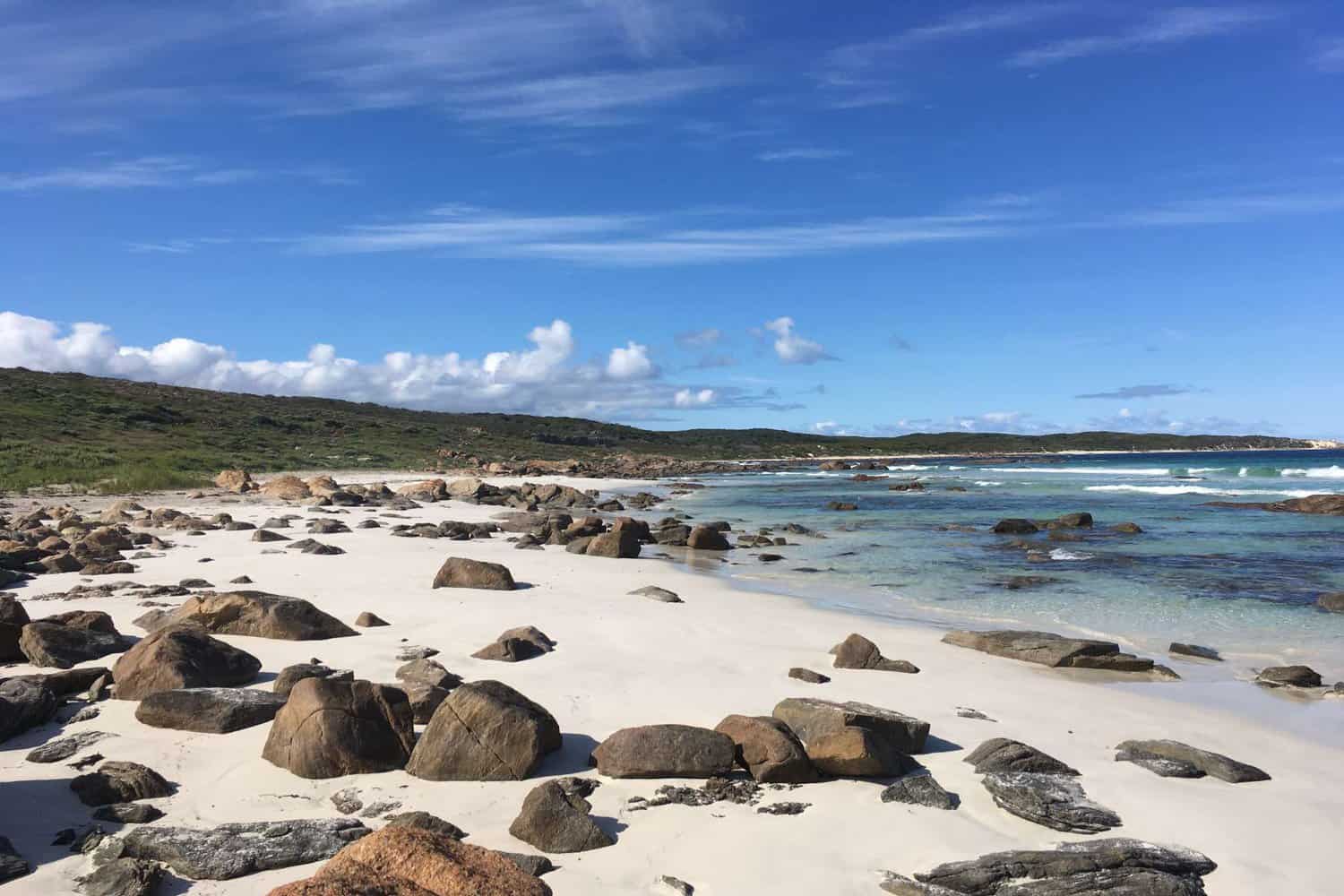 Scattered rocks and clear, shallow waters edge the white sands of a secluded Margaret River beach, set against a backdrop of lush greenery and a bright blue sky dotted with fluffy clouds.