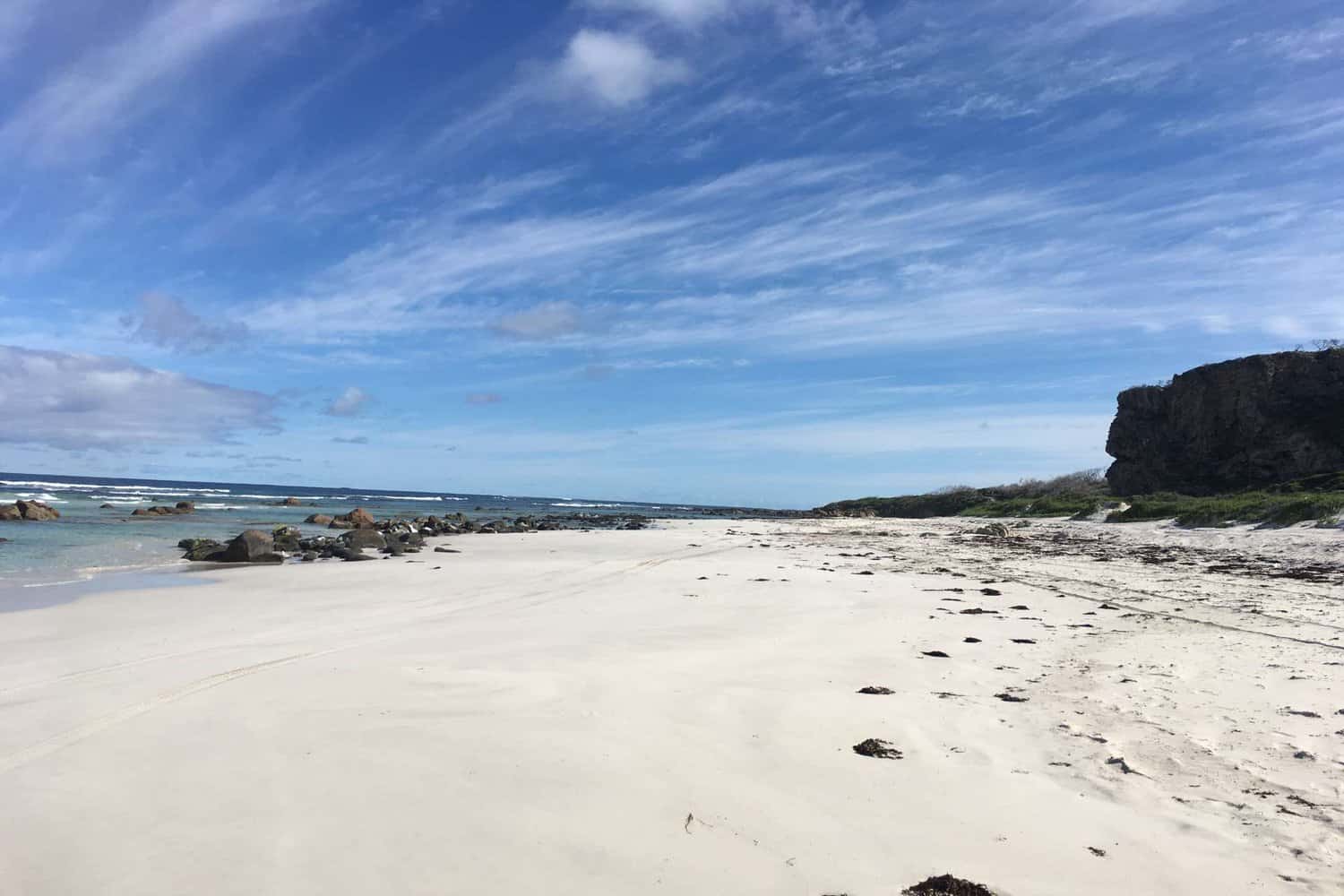 Expansive white sandy beach in Margaret River with scattered rocks leading to the ocean, under a striking blue sky with wispy clouds.