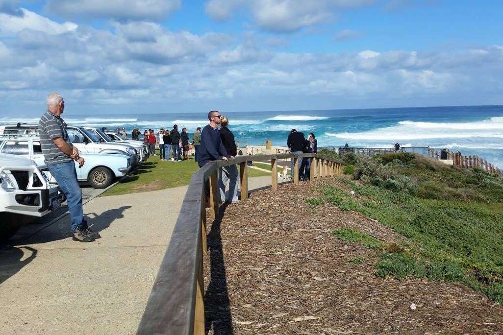 Spectators gather along a wooden fence at a viewpoint overlooking the dynamic surf of Margaret River beaches, with a clear blue sky above and vehicles parked nearby, highlighting a popular spot for ocean watching.