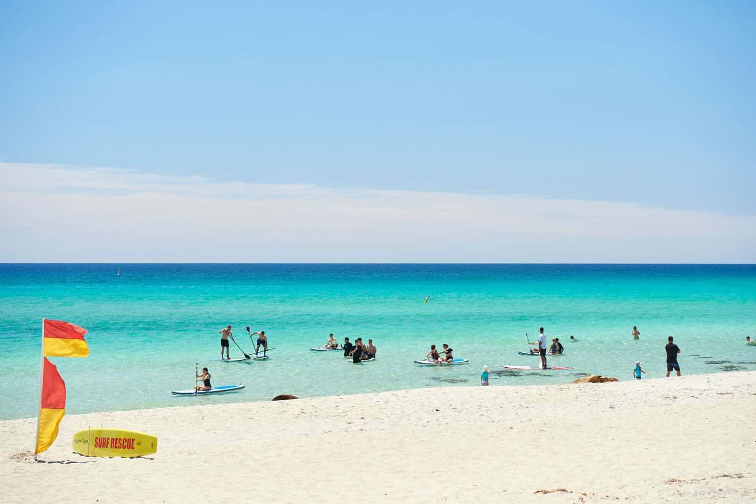 Vibrant beach day at Dunsborough with clear turquoise waters, people paddleboarding, and a surf rescue sign in the foreground.