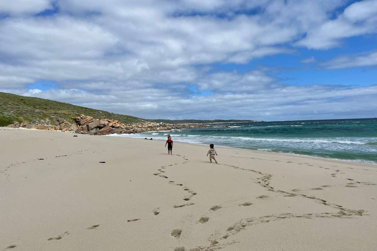 Footprints trace the journey of beachgoers on the soft, golden sands of a Margaret River beach, with the ocean's turquoise waves gently approaching the shore and a rugged hillside framing the scene