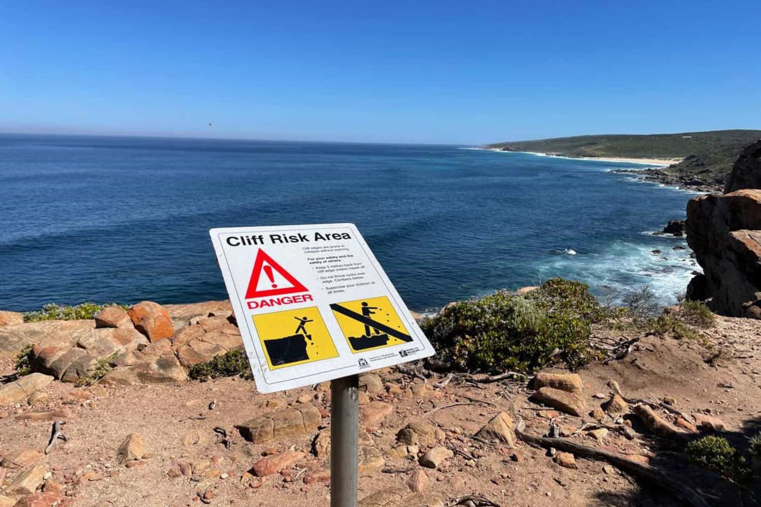 Warning sign alerting to a 'Cliff Risk Area' with symbols indicating the danger of falling rocks and cliff edges, placed against the backdrop of a stunning coastal view at Wilyabrup Cliffs with clear blue waters.