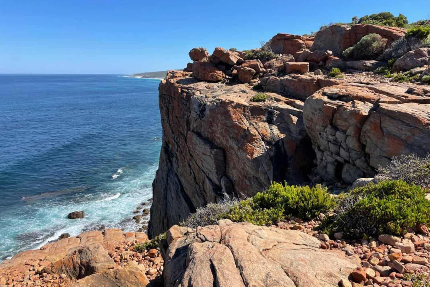 Scenic view of the rugged Wilyabrup sea cliffs along the coastline, with layered rock formations and shrubs overlooking the azure ocean waves, under a clear blue sky.