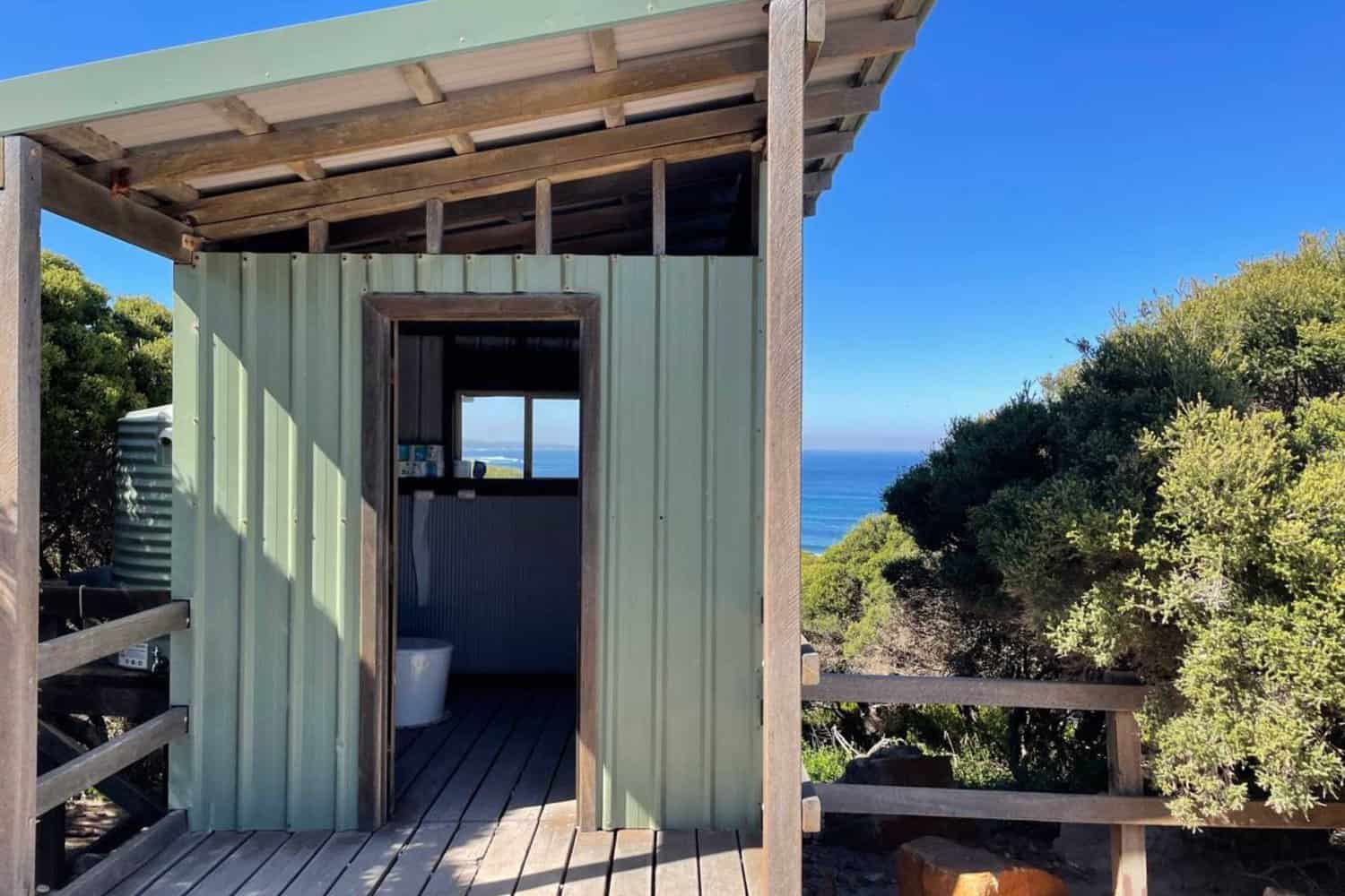 A small green coastal outhouse with an open window offering a view of the Indian Ocean, nestled among native shrubbery near the Wilyabrup Sea Cliffs.