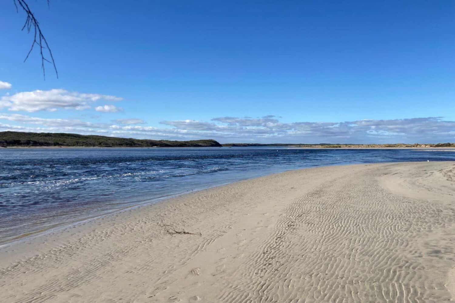 A tranquil, sandy river mouth opening into the ocean at a dog-friendly beach near Margaret River, with clear blue skies and distant headlands.