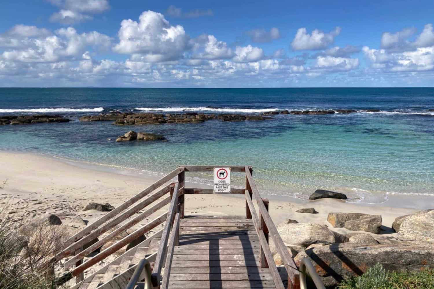 A wooden staircase leads down to the inviting clear waters of an Augusta beach, Granny's Pool, framed by rock formations that create natural pools, with a 'No Dogs' sign reminding visitors of the sanctuary's rules under a sky dotted with fluffy clouds.