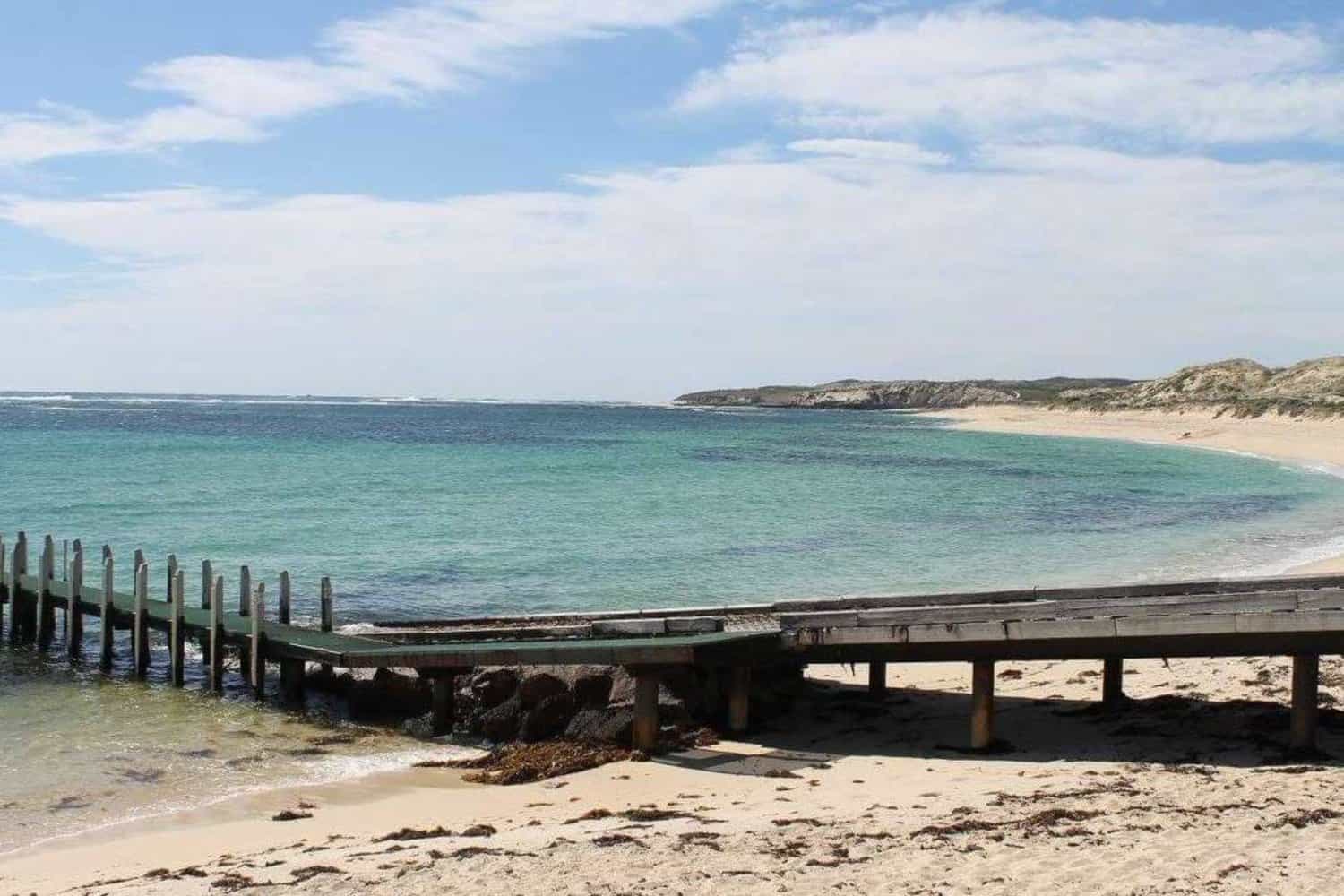 Old wooden jetty reaching out into the clear teal waters of a dog-friendly beach near Margaret River, with untouched sand dunes under a partly cloudy sky.