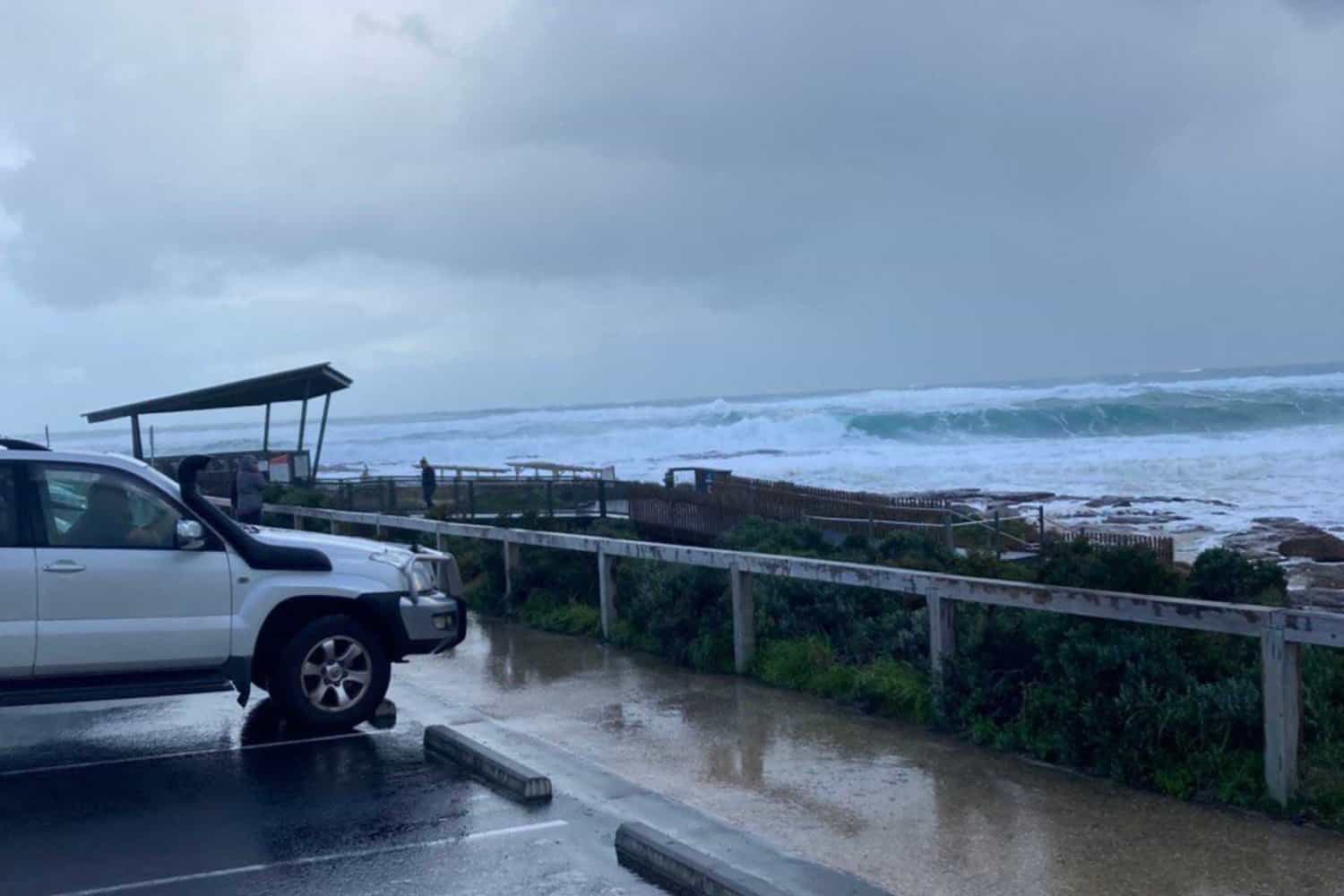 A white SUV parked by a coastal lookout on a stormy day, with turbulent ocean waves in the background, capturing a dramatic and moody seascape along the Australian coast.