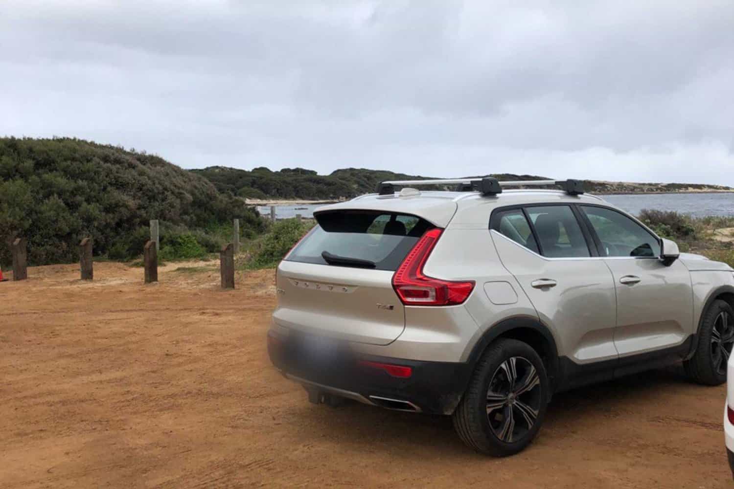 A silver Volvo XC40 parked on a sandy lot near a coastal area, with dense shrubbery and a glimpse of calm sea waters in the background, typical of a serene beachside parking area in Australia.