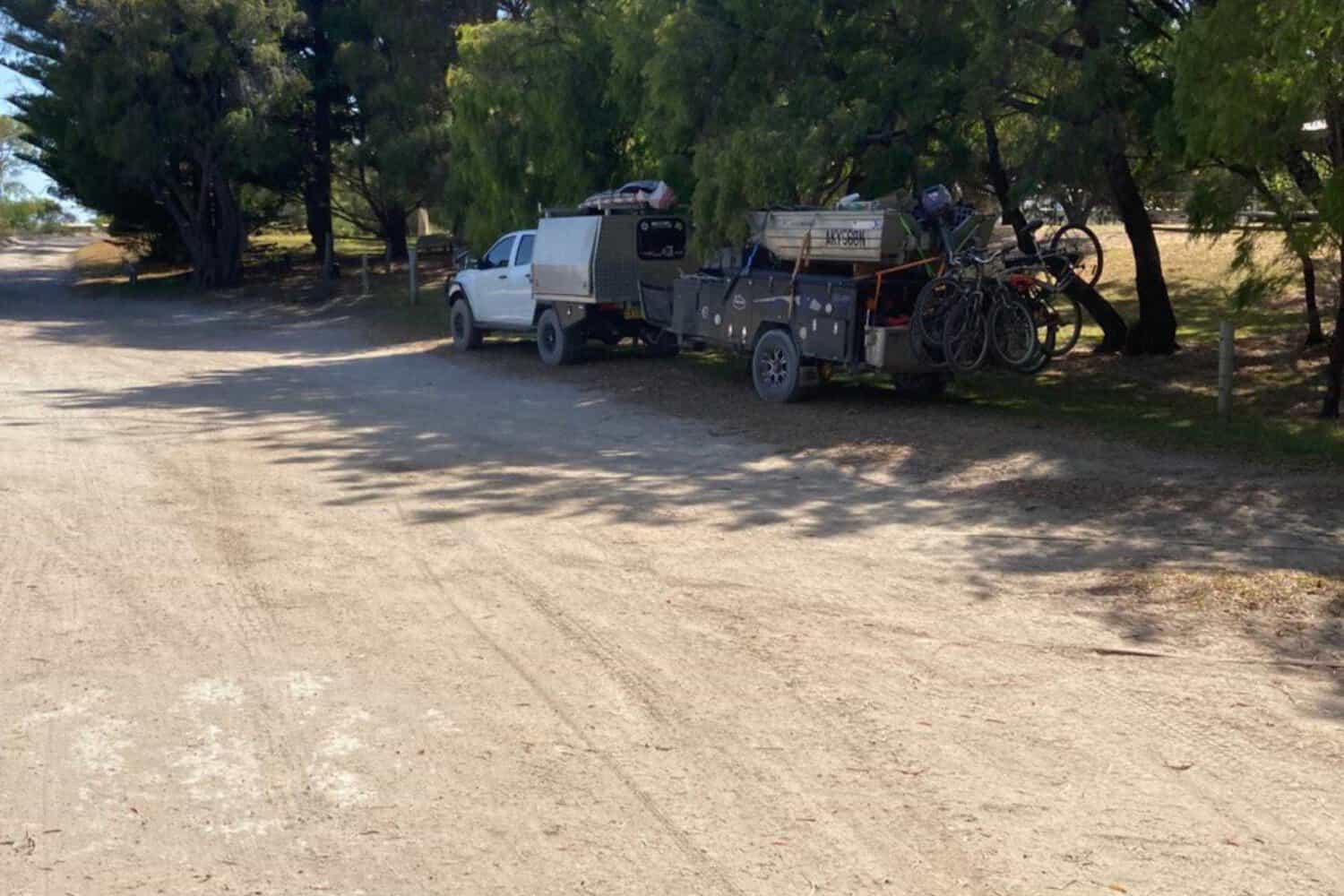 A white pickup truck with a camper trailer and bicycles parked under the shade of trees on a dusty road, suggesting preparations for an outdoor adventure in the Margaret River region.