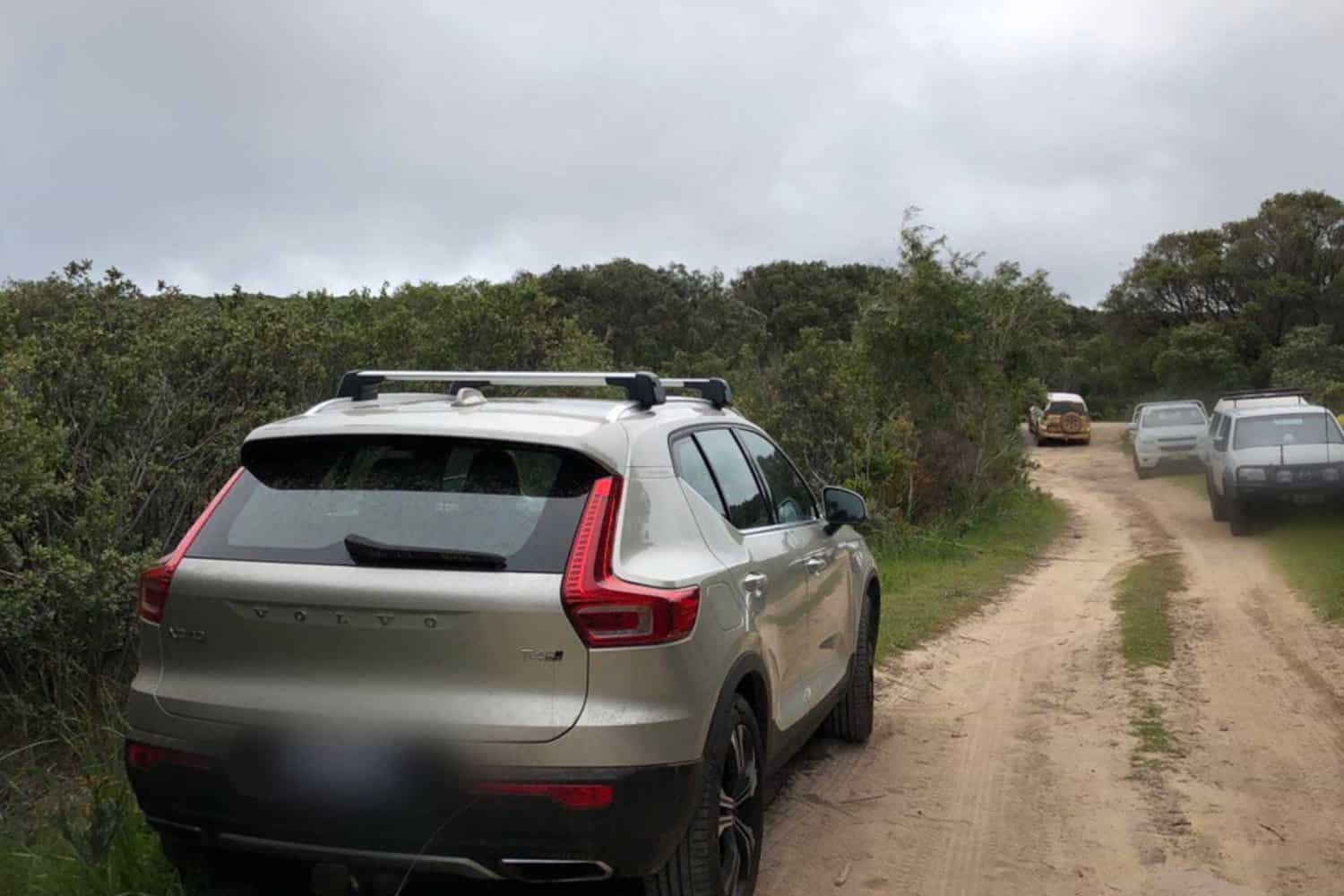 A line of vehicles, led by a silver Volvo XC40, navigates a narrow dirt road surrounded by dense green shrubbery under an overcast sky. The focus is on the Volvo's rear, highlighting its modern design and the rugged, natural setting of an outdoor trail.