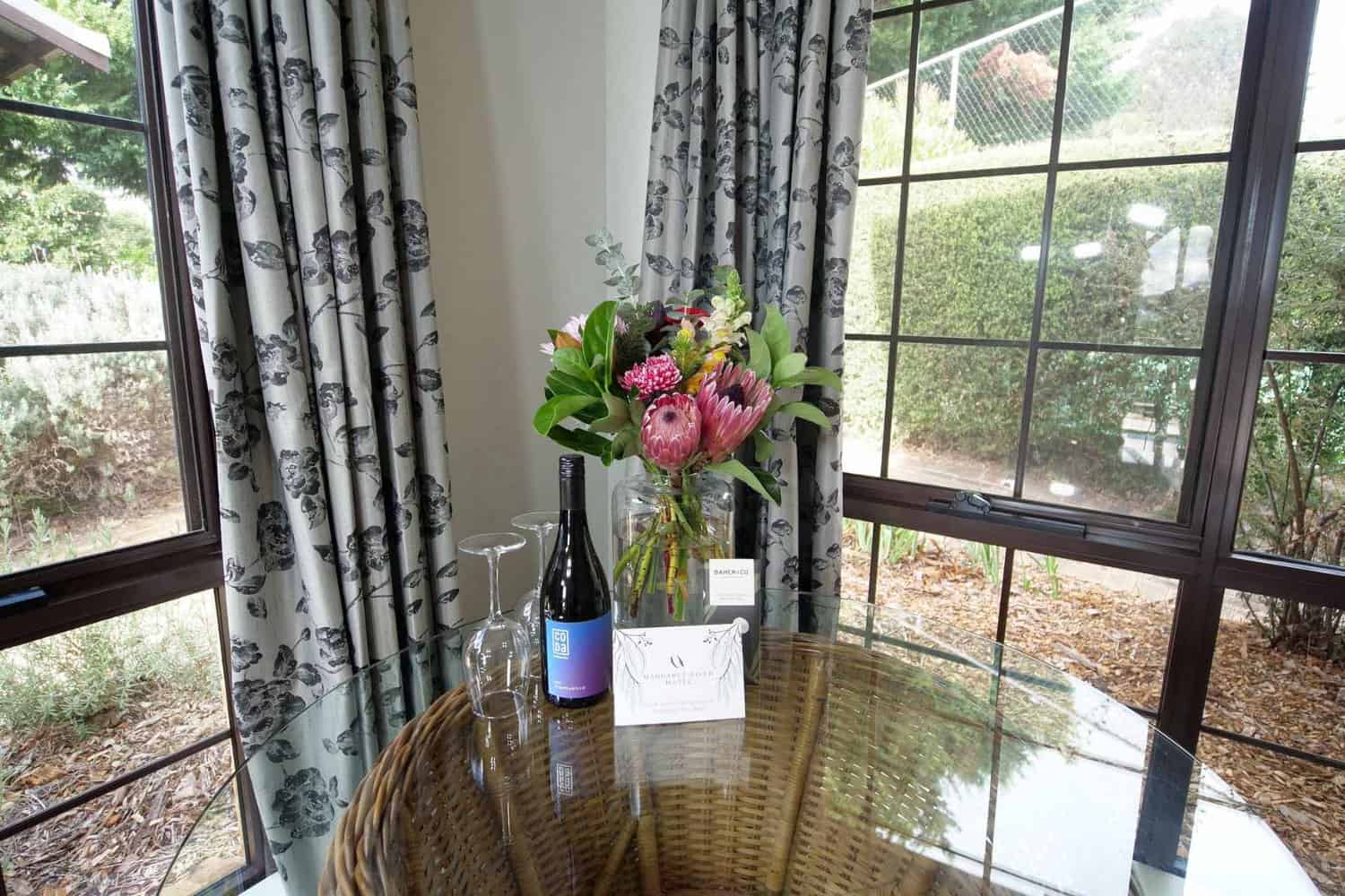Elegant hotel room detail with a bottle of wine and two glasses on a glass-topped table, complemented by a vibrant bouquet of flowers, offering a welcoming atmosphere next to a window with garden views.