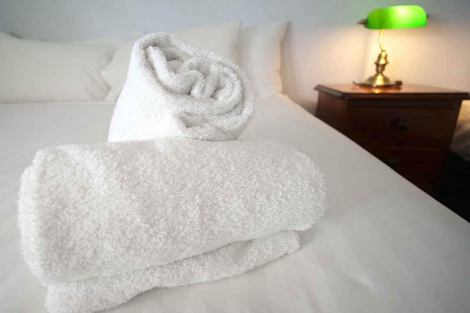 Close-up of fluffy white towels on a hotel bed with crisp linen, with a vintage nightstand and green lampshade in the background, suggesting a cozy and welcoming guest room.