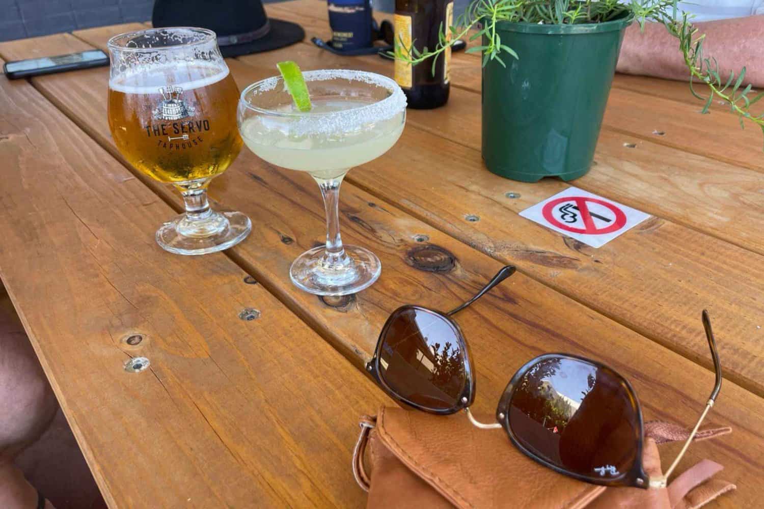 A photo of a pint of beer and a handcrafted cocktail on a wooden table at a local Margaret River brewery. The beer has a thick creamy head, and the cocktail is garnished with fruit and herbs. The rustic wooden table with small plant as the centrepiece evoke a warm and cozy atmosphere, inviting you to sit and enjoy a drink with friends.