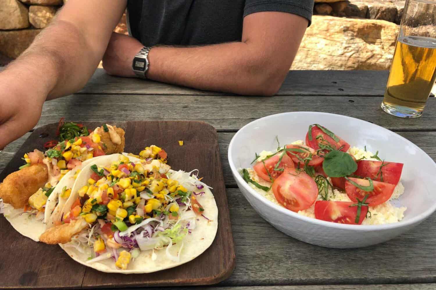 A delectable dish of fish tacos and fresh tomatoes served on a wooden table at a Margaret River brewery, with a pint of beer on the side. The tacos are garnished with a corn salsa, and the side of fresh tomatoes add a bright pop of color to the table.