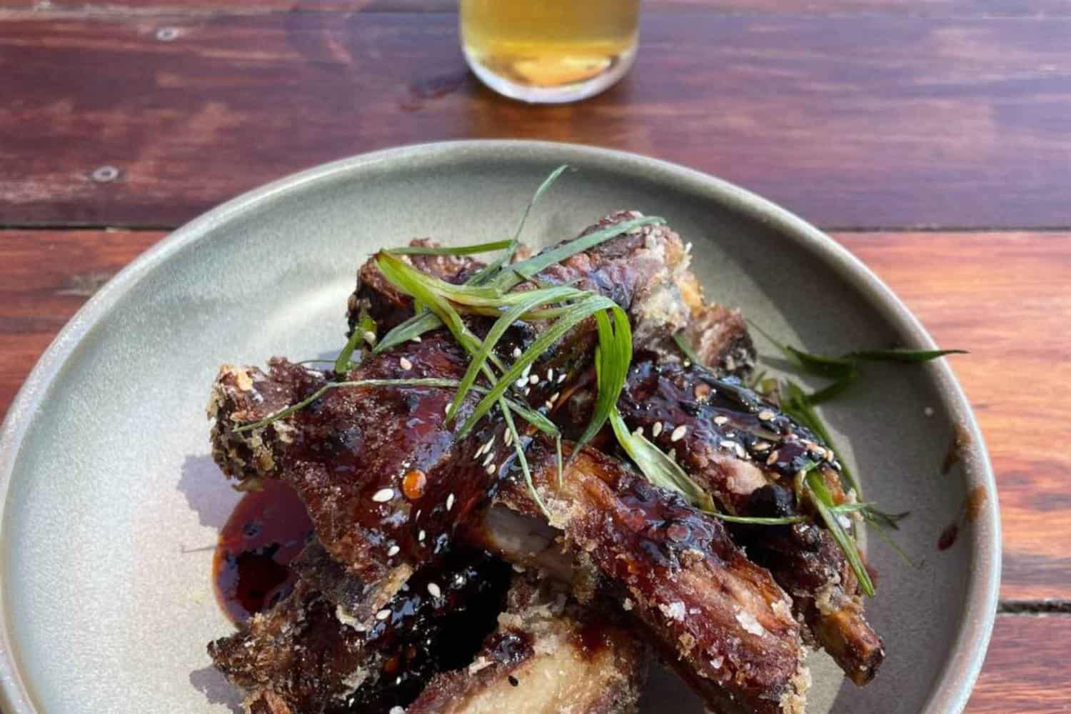 Succulent glazed ribs garnished with green onions on a ceramic plate, served on a rustic wooden table with a glass of beer in the background, showcasing a restaurant's specialized dish.