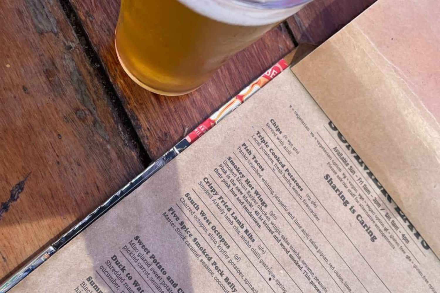 A menu at the Beerfarm Margaret River displayed on a wooden table accompanied by a glass of beer. The menu features a variety of dishes, ranging from burgers and fries to share plates and desserts. 
