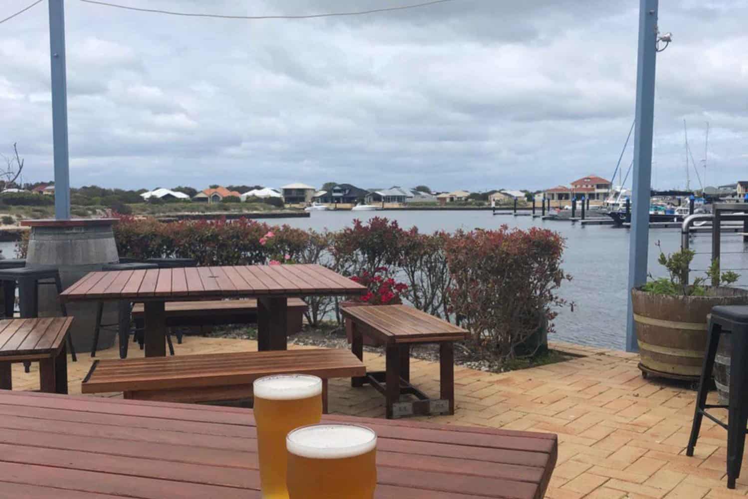 A photo of frosty beers on a wooden table at a brewery overlooking the water. The serene view of the water adds to the relaxing vibe of the brewery, perfect for enjoying a cold pint and the company of friends. The view creates a cozy atmosphere that invites you to stay awhile and take in the beautiful surroundings.
