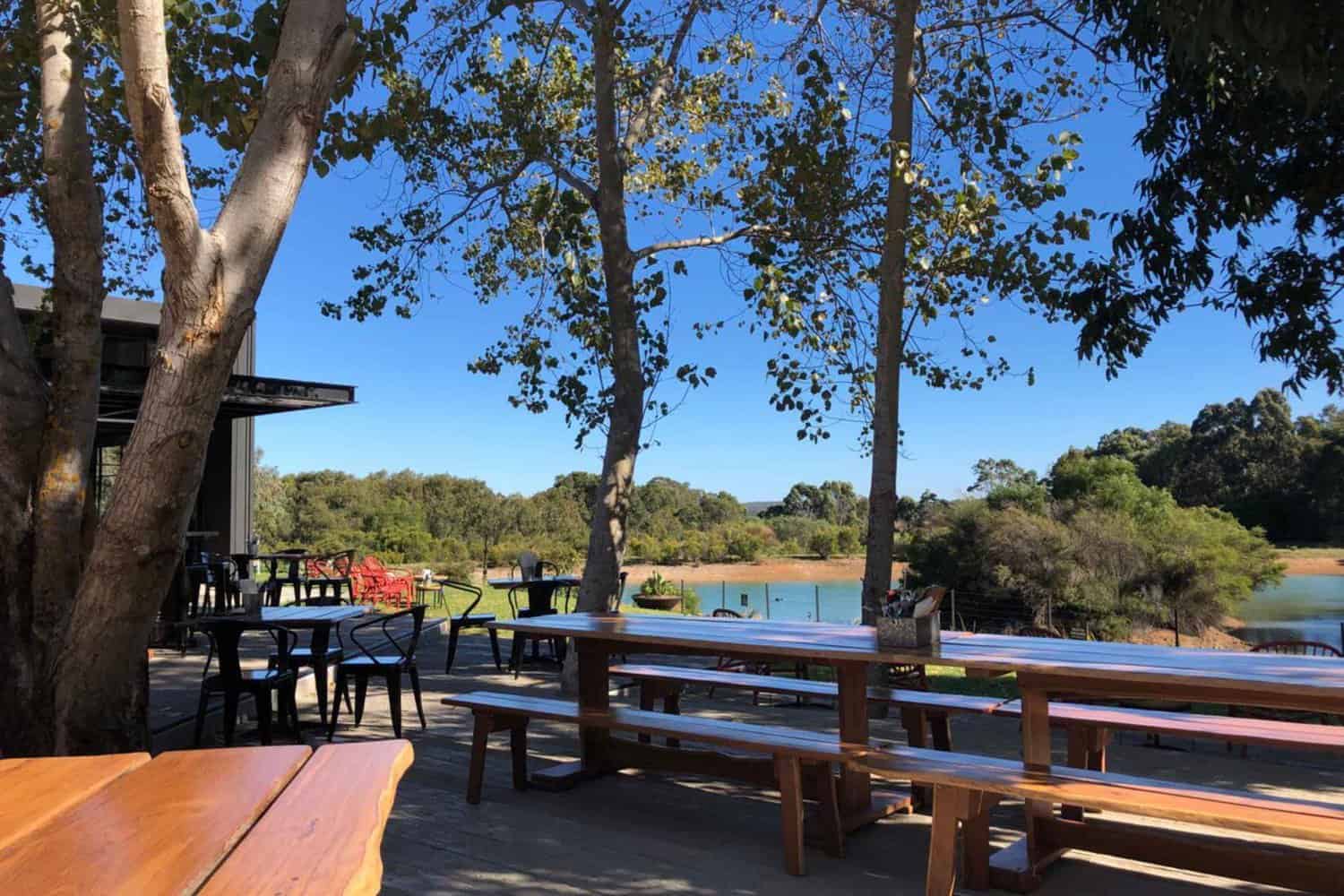 A serene view from a Margaret River Brewery, looking over the outdoor tables onto a peaceful lake with Australian bush in the distance. The lake is surrounded by trees, creating a tranquil and secluded atmosphere.
