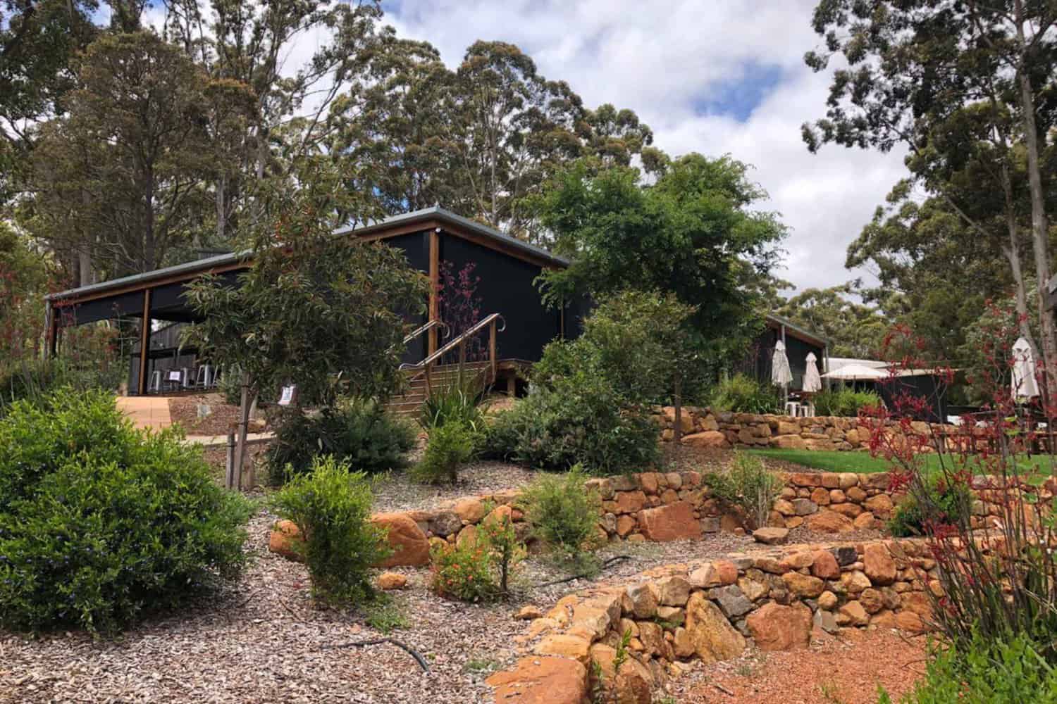 Building facade of the Margaret River Brewhouse, featuring a wooden exterior with large glass windows, outdoor seating, and shrubs, with trees in the background