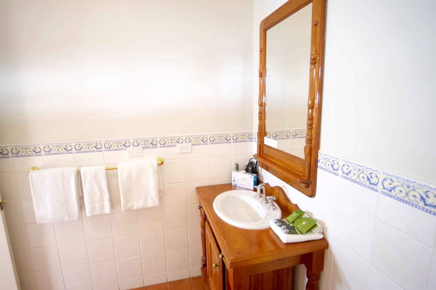 A bright, clean bathroom with white walls and a wooden vanity that holds a sink with a silver faucet. Above the vanity is a large wooden-framed mirror, and on the wall, there is a row of decorative blue and white tiles. White towels hang neatly on a rail to the left of the vanity.