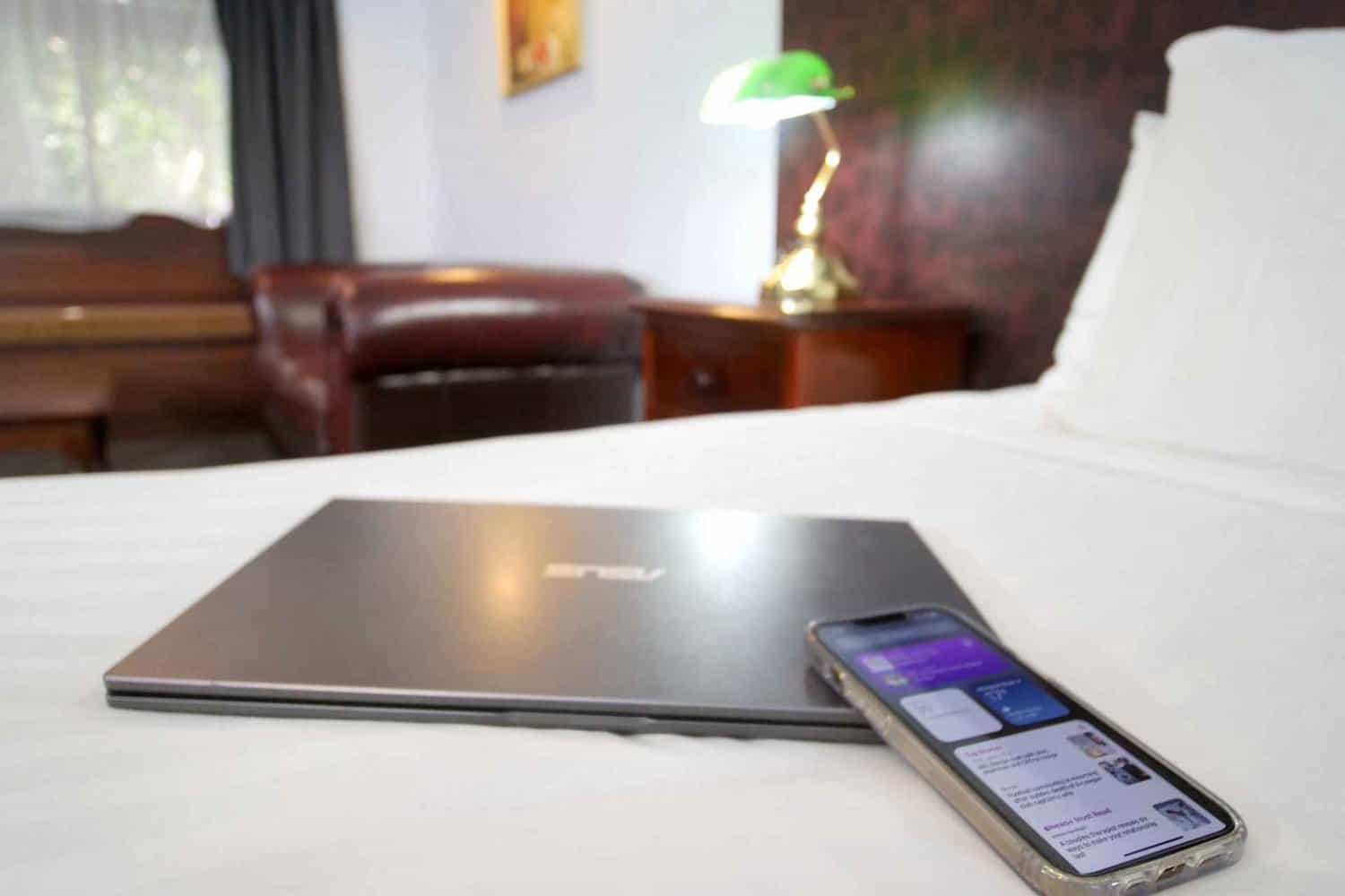 A close-up view of a laptop and phone on a hotel bed, with a couch and bedside table in the distance, signifying a comfortable environment for guests to relax after securing their hotel deposit.