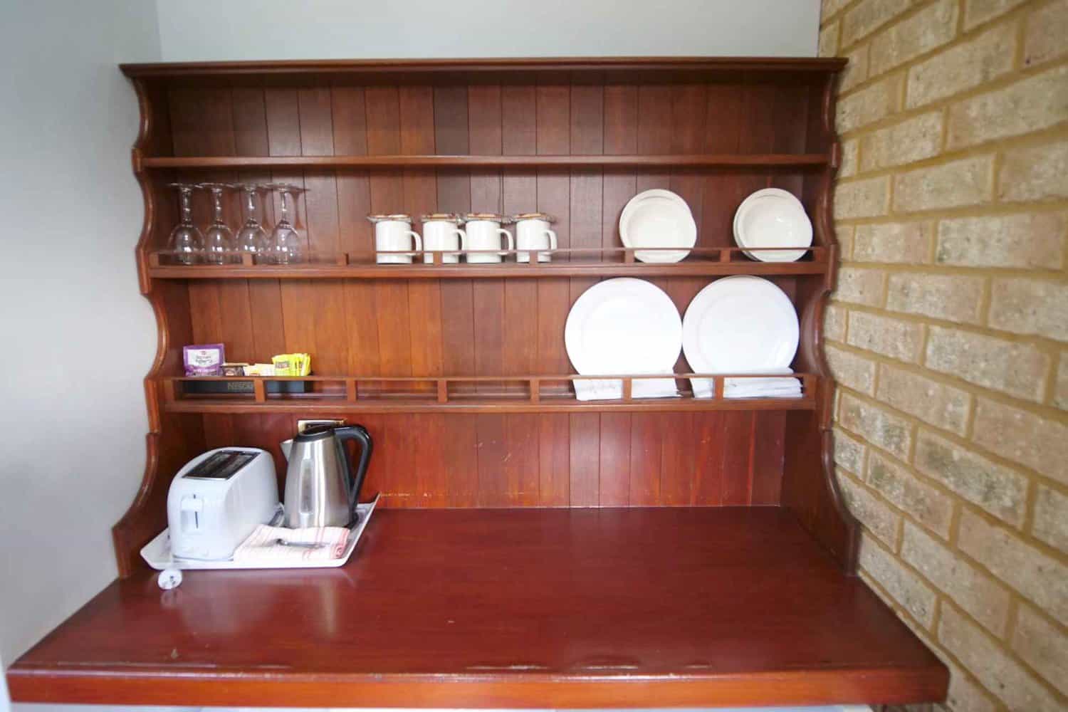 Hotel room amenities with a wooden shelf displaying a tea set, white plates, and glasses, alongside an electric kettle and toaster, ready for a comfortable stay.