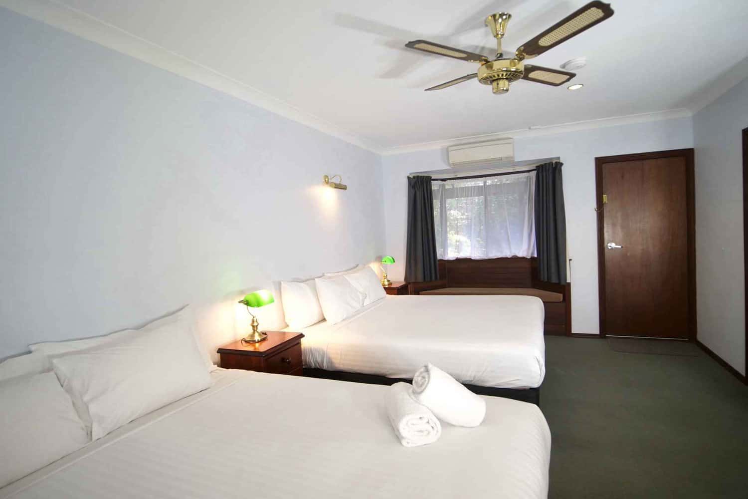 A twin hotel room with two beds, crisp white bedding, and green shaded bedside lamps, complete with a ceiling fan and a view of the window drapes partially closed.