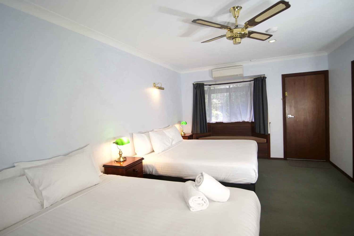 A spacious family room in a hotel with two king-size beds, sunlight streaming through the window, bedside tables with lamps, and towels on the bed, providing a comfortable stay for guests who have made their hotel deposit.