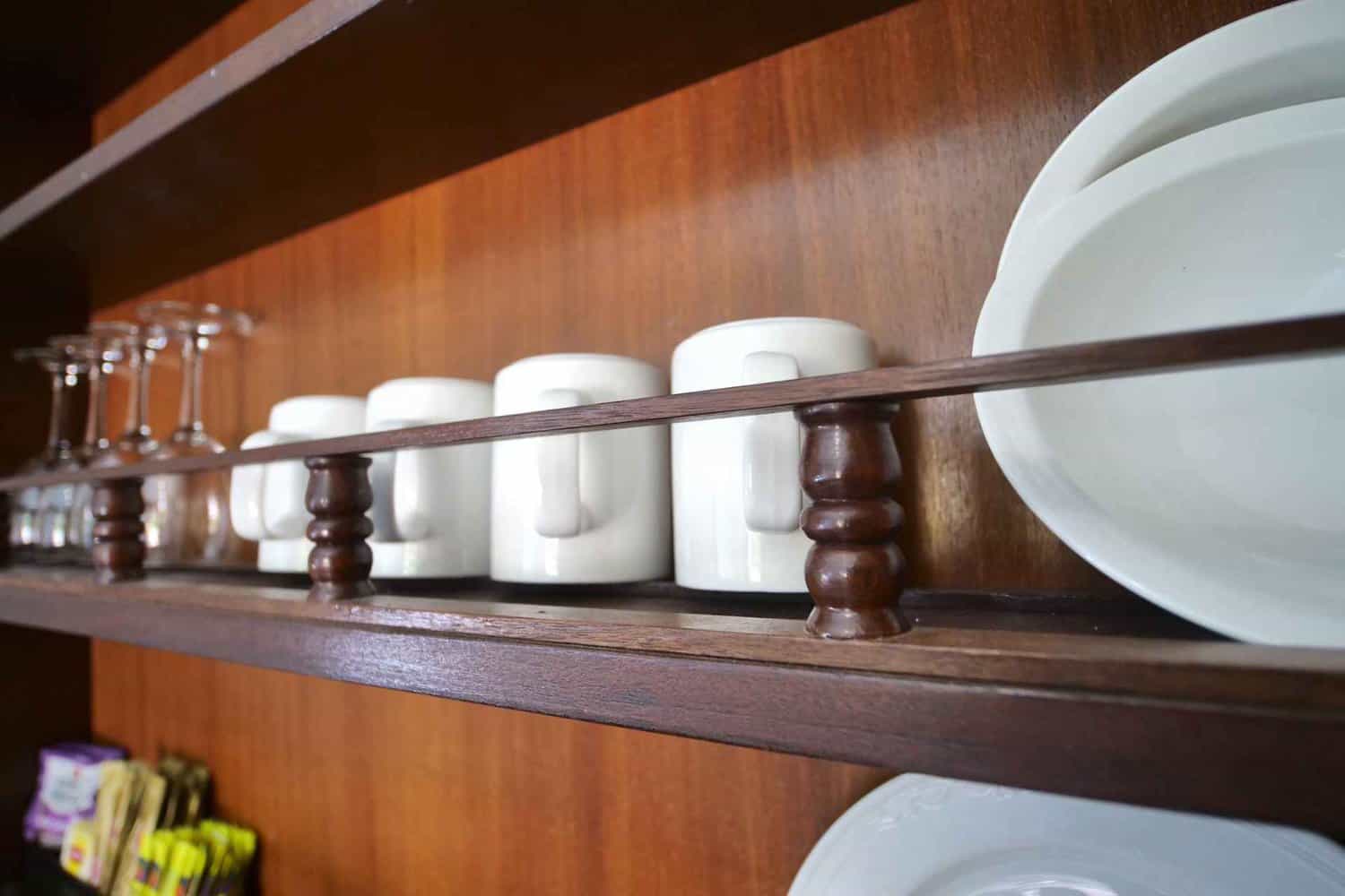 Close-up of a wooden shelf in a hotel room, neatly arranged with white porcelain coffee mugs and plates, alongside clear glass stemware, set against a rich, dark wood background.