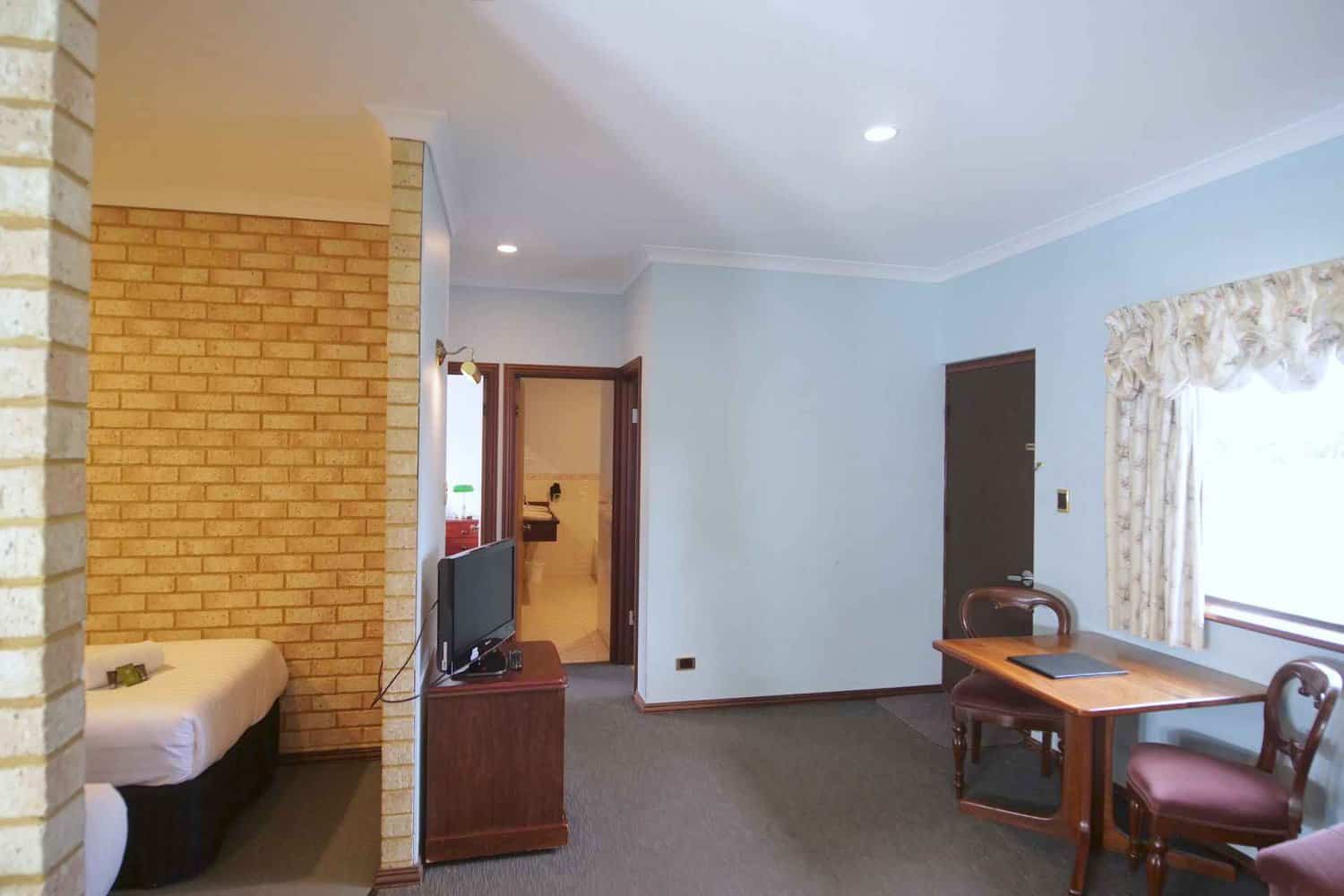 Bright and airy hotel suite featuring a sleeping area with a single bed, a flat-screen TV on a wooden stand, and a work desk with chairs, showcasing a comfortable and functional space for guests.