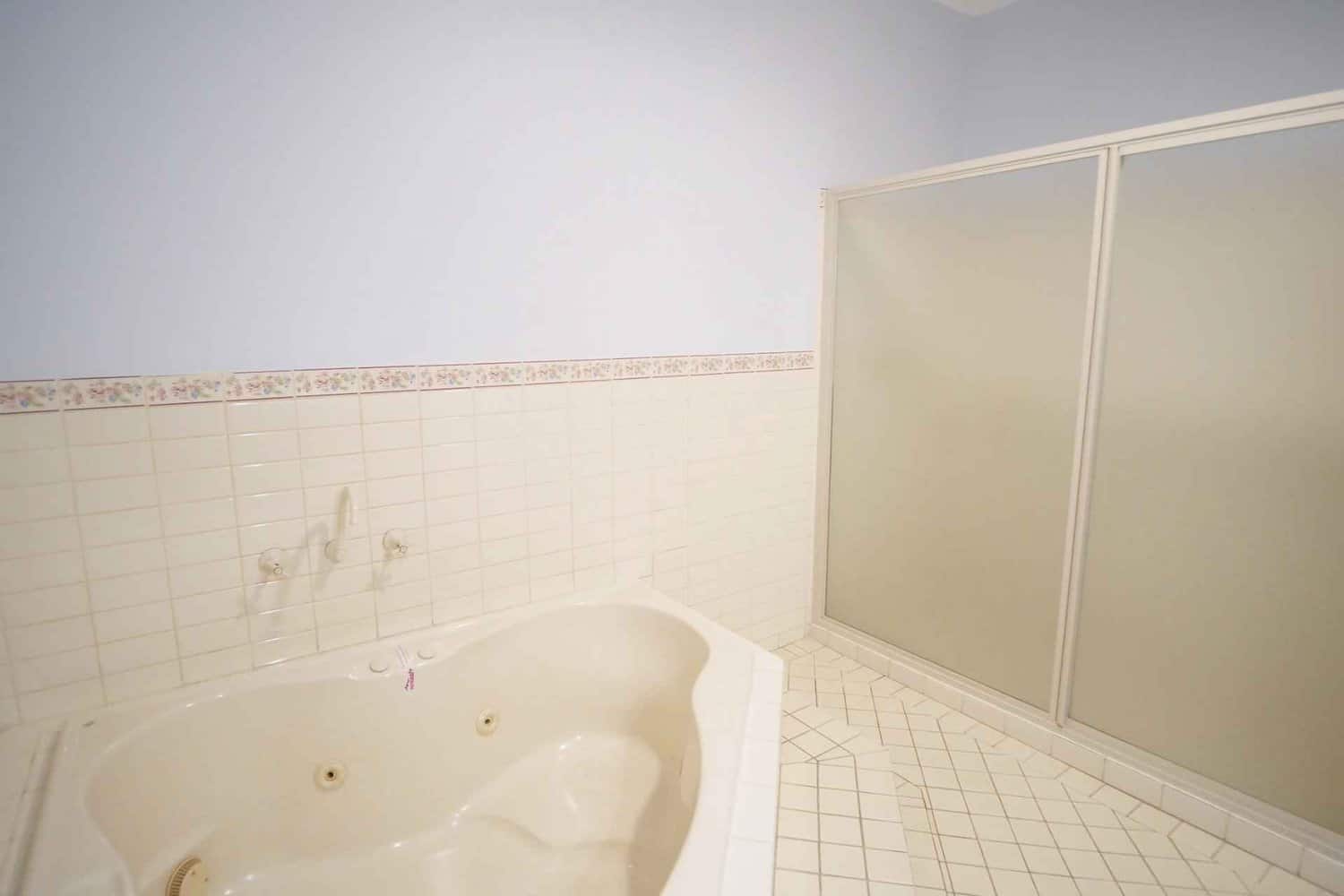 Bright and clean hotel bathroom featuring a large corner jacuzzi tub next to a spacious shower area, with white tiled walls and floral border detailing.