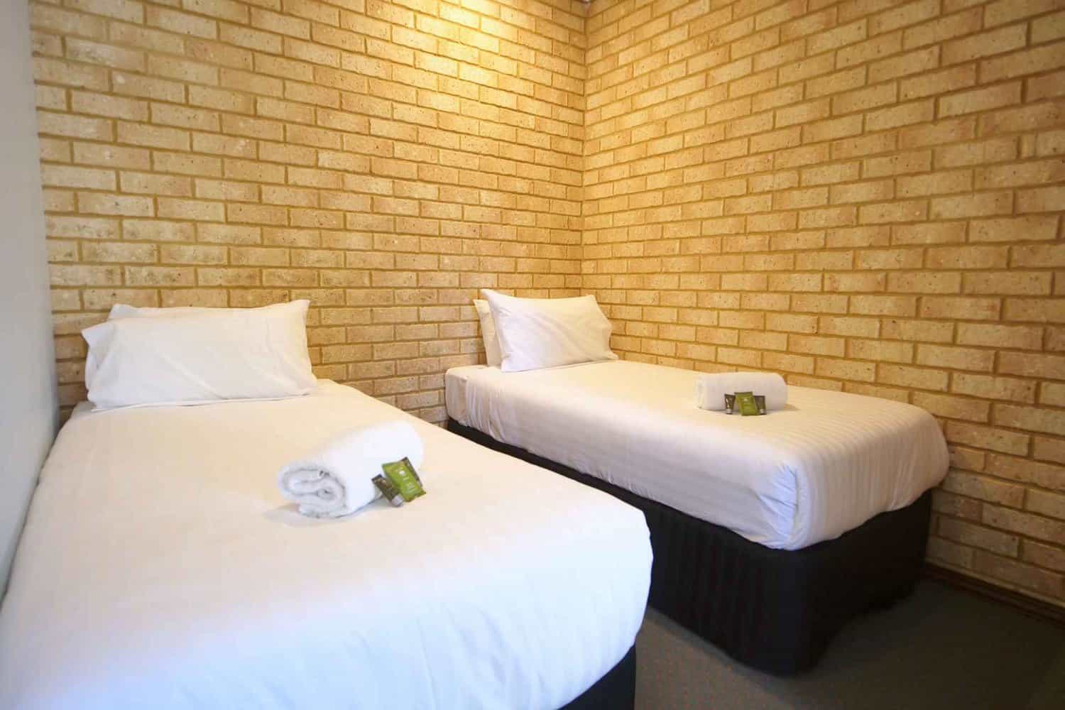 Twin single beds with white linens in a hotel room featuring an exposed brick wall, each bed neatly made and accompanied by a small green amenity pack, offering a simple yet comfortable accommodation.