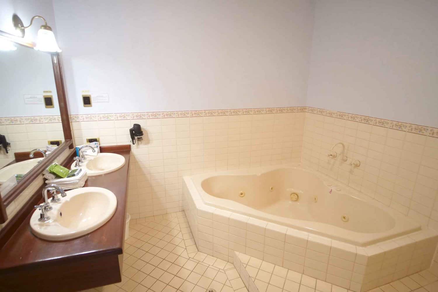 Luxurious hotel bathroom boasting a relaxing corner spa, an elegant vanity with double sinks, a large mirror over the sinks, and fresh towels accompanied by a selection of bathroom amenities, enhancing the comfort and pampering experience during extended stays