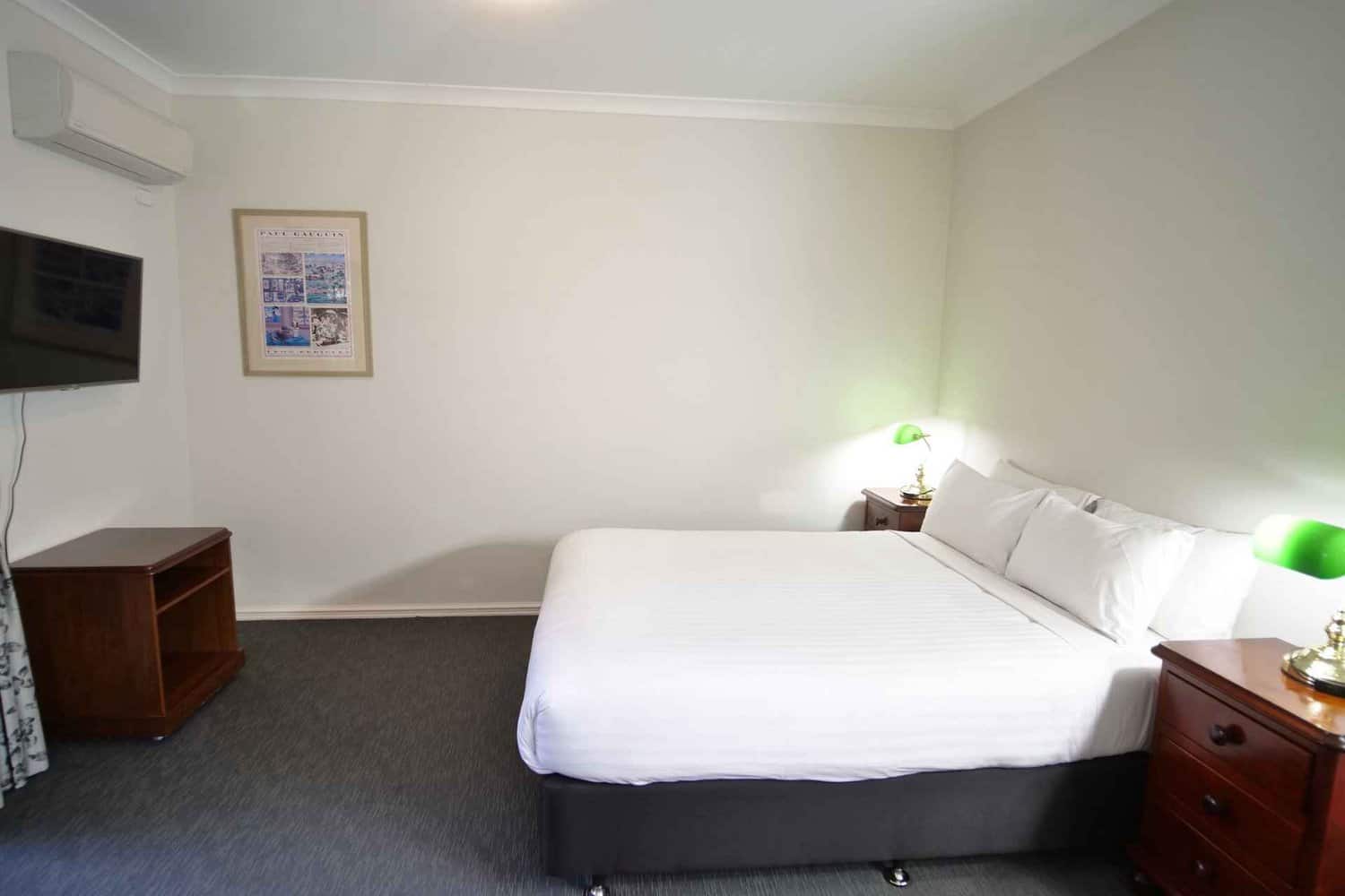 Spacious hotel room featuring a comfortable queen-size bed, flanked by bedside tables with lamps, a wall-mounted TV for entertainment, air conditioning for climate control, and an eye-catching painting on the wall, creating a relaxing atmosphere for guests during their stay