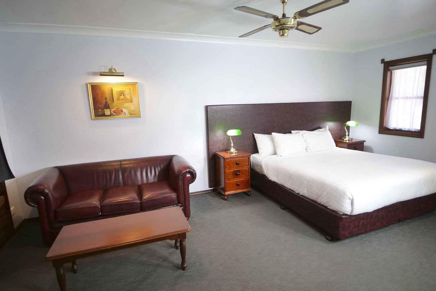 Elegant hotel room featuring a plush king-size bed with an imitation leather bedhead, bedside tables with lamps for cozy lighting, a comfortable couch for lounging, and a tasteful painting of a wine bottle on the wall, creating a warm and inviting atmosphere for guests
