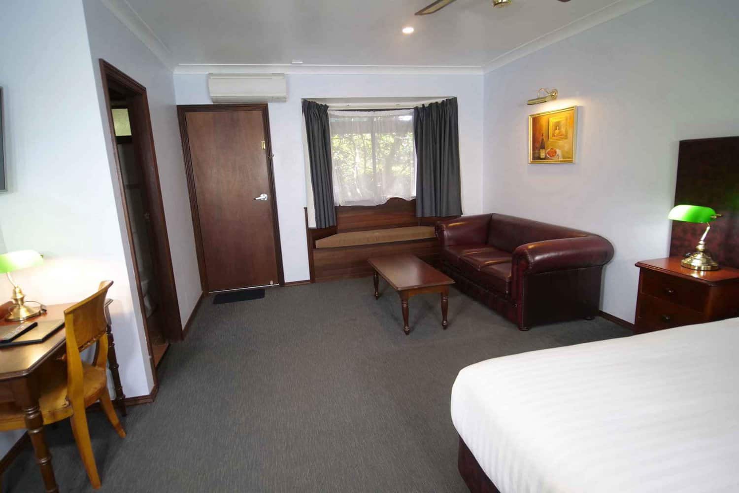 Elegant hotel room featuring a plush king-size bed with an imitation leather bedhead, bedside tables with lamps for cozy lighting, a comfortable couch for lounging, and a tasteful painting of a wine bottle on the wall, creating a warm and inviting atmosphere for guests