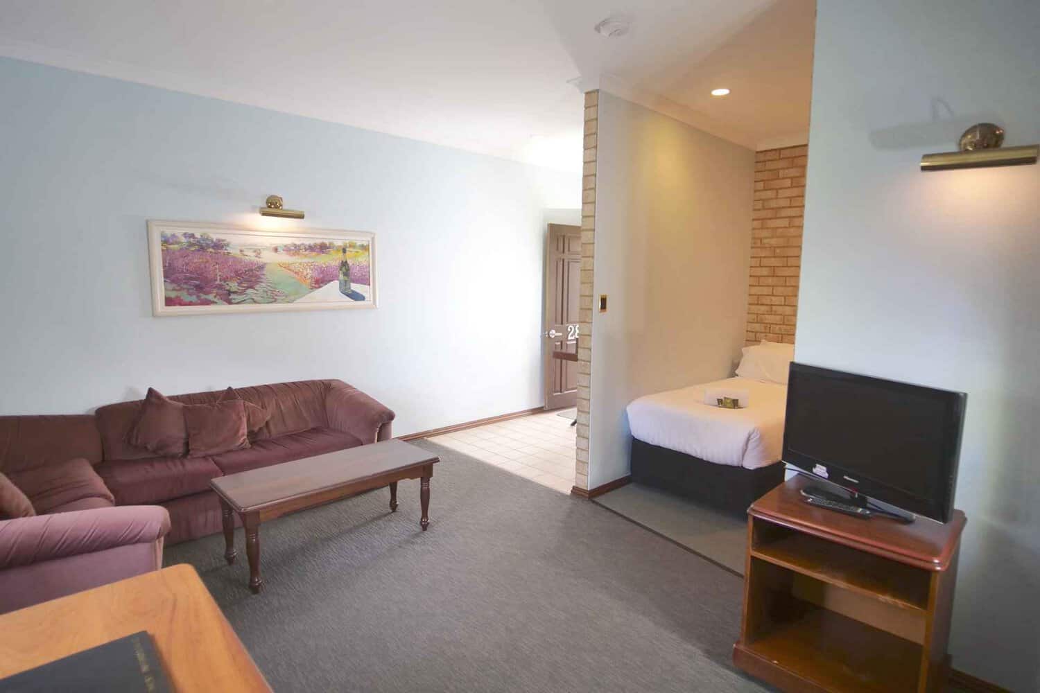 Inviting hotel loungeroom featuring cozy couches, a coffee table, television for entertainment, and a single bed, with an open door leading to additional spaces, complemented by a beautiful painting of a vineyard on the wall, creating a welcoming atmosphere for extended stays