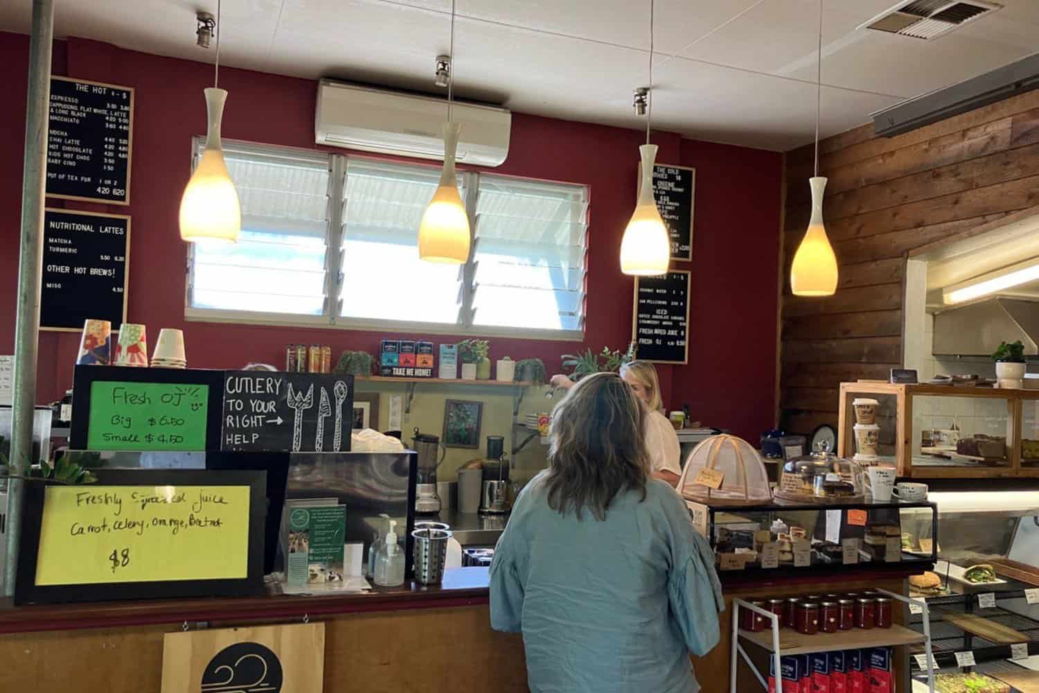 A bustling coffee shop in Margaret River, featuring a lady ordering the best coffee from the counter, an appetizing display of cakes and sandwiches, informative signs on the wall, and a professional coffee machine ready to brew the perfect cup.