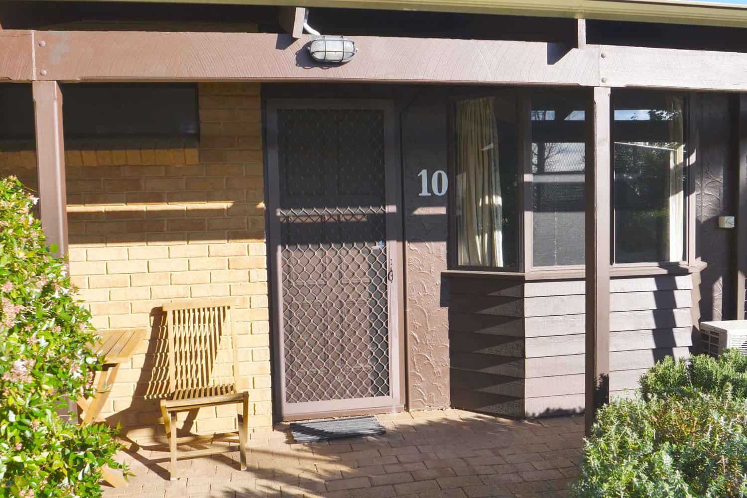 Exterior view of a motel room entrance featuring a numbered door with a security screen, a wooden bench, and surrounding greenery.